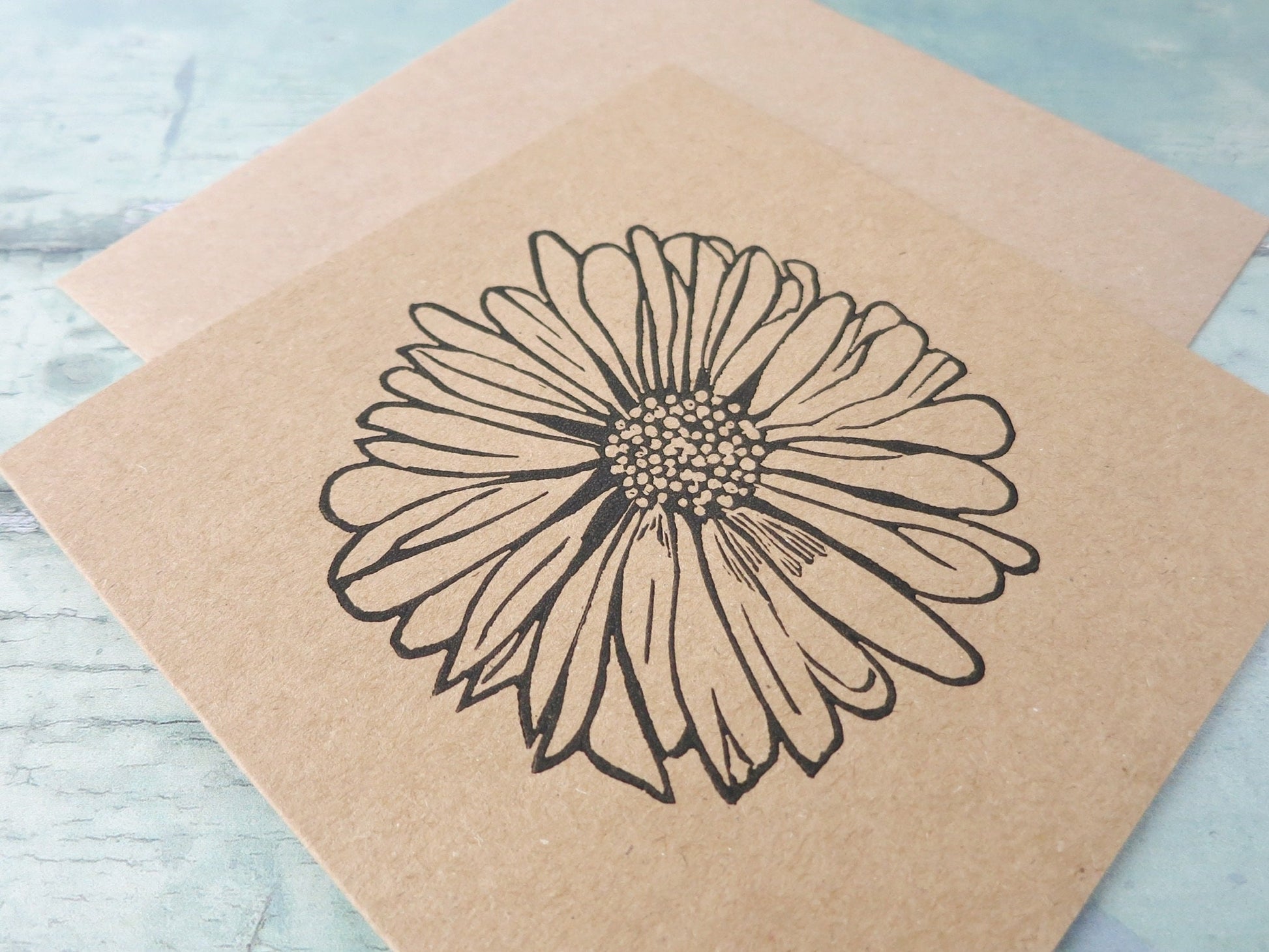 Close up of Kraft paper card with an original daisy lino print laid on a textured surface. 