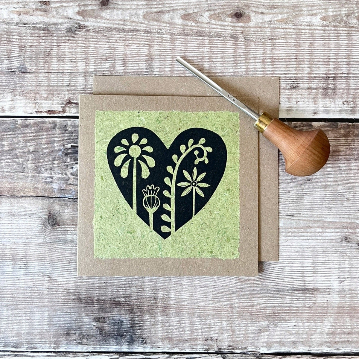 Square kraft greeting card with a black floral heart lino print on pale green handmade paper mounted on the front. Laid on a wooden background. Photo includes the lino cutting tool used.