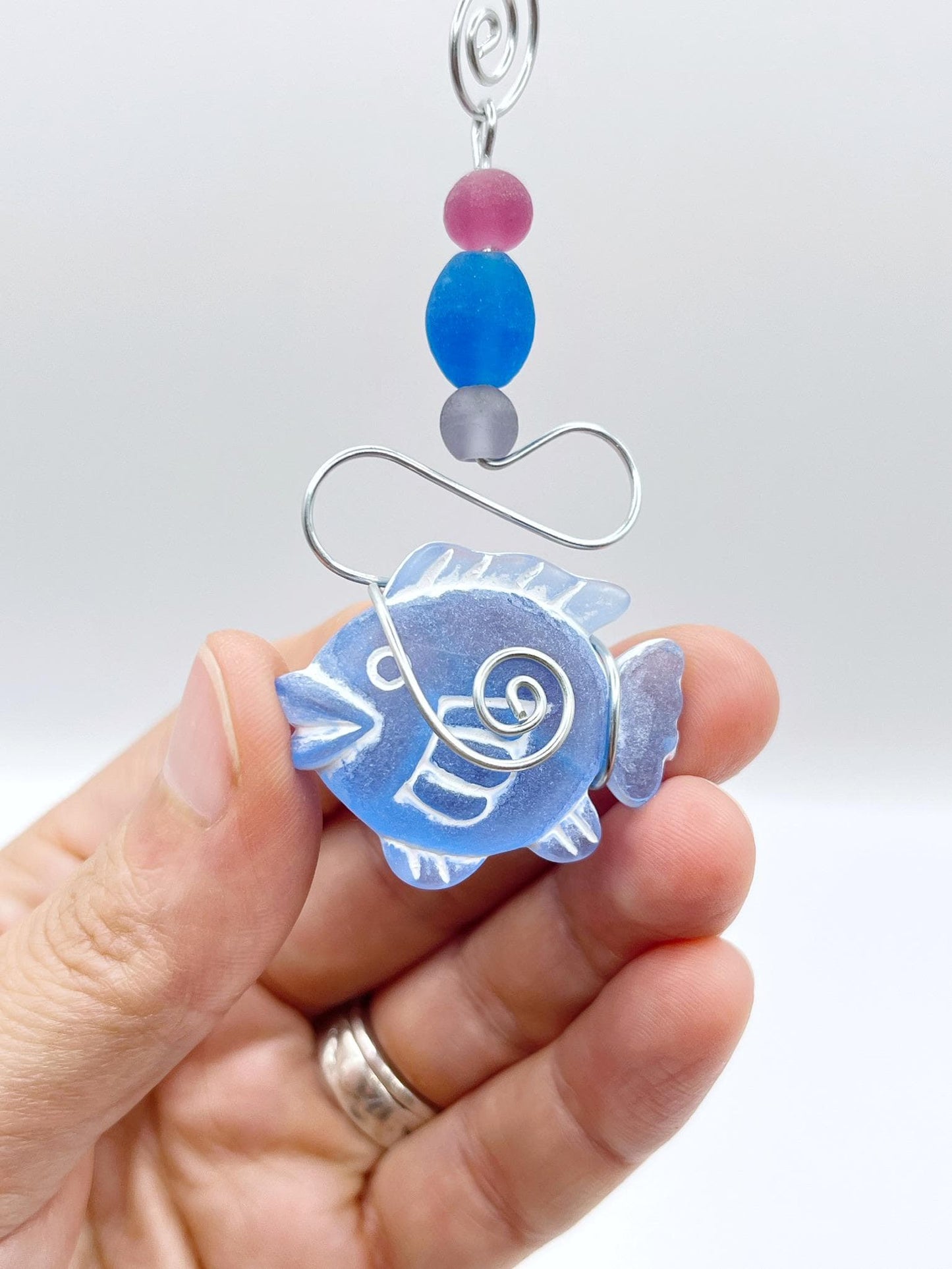Decorative hanging ornament with blue fish design, wire and colourful beads, held in a hand against a white background
