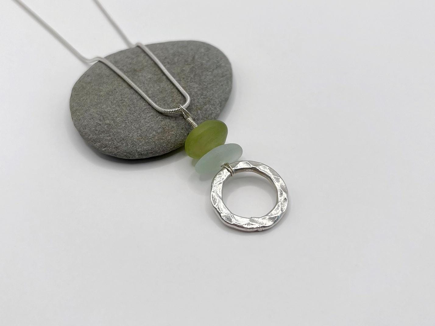 Necklace with a silver ring pendant and green jade and sea glass laid  on a grey stone and white background