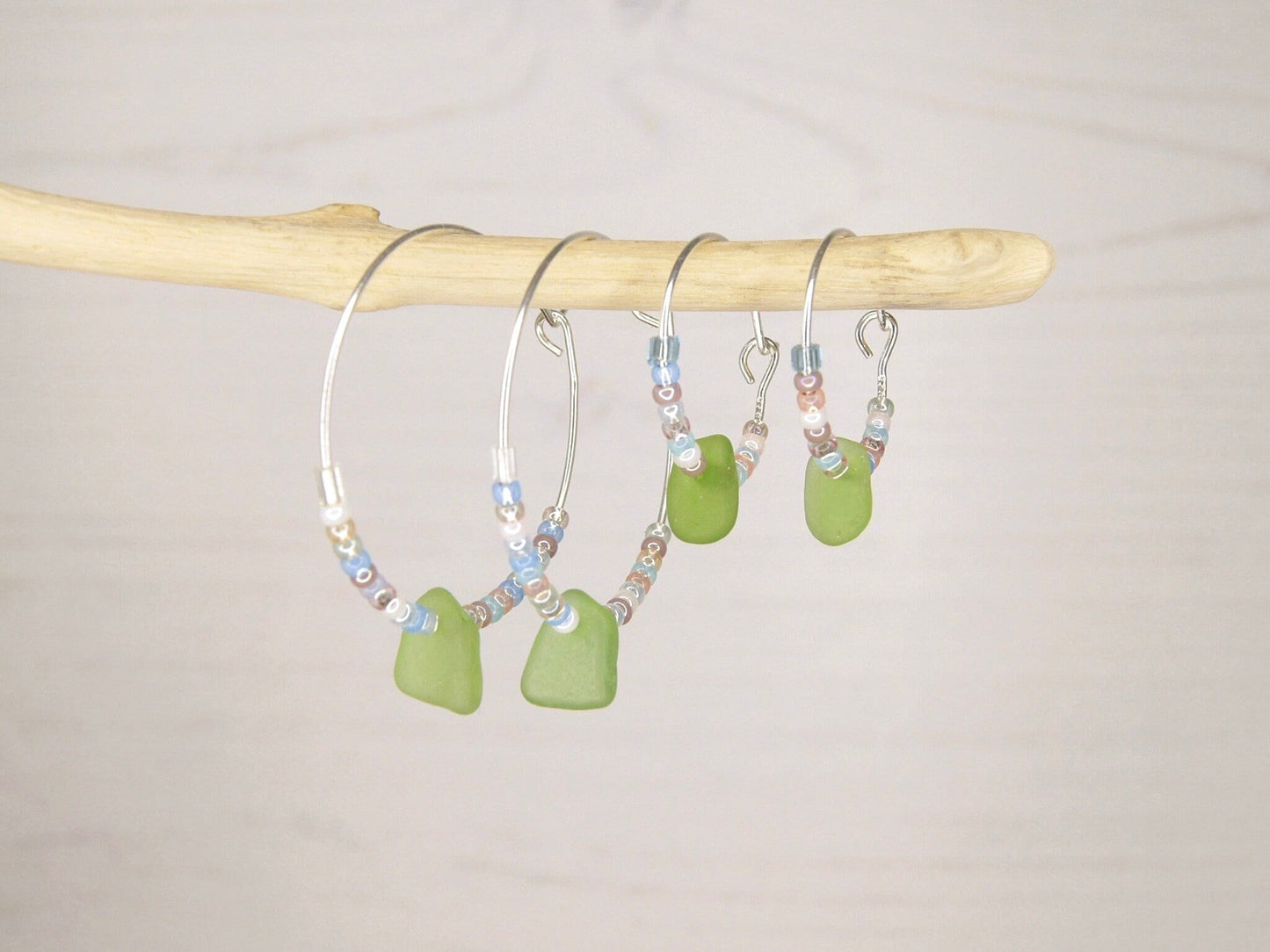 Two pairs of hoop earrings with pastel seed beads and green sea glass hanging on a driftwood against a plain background