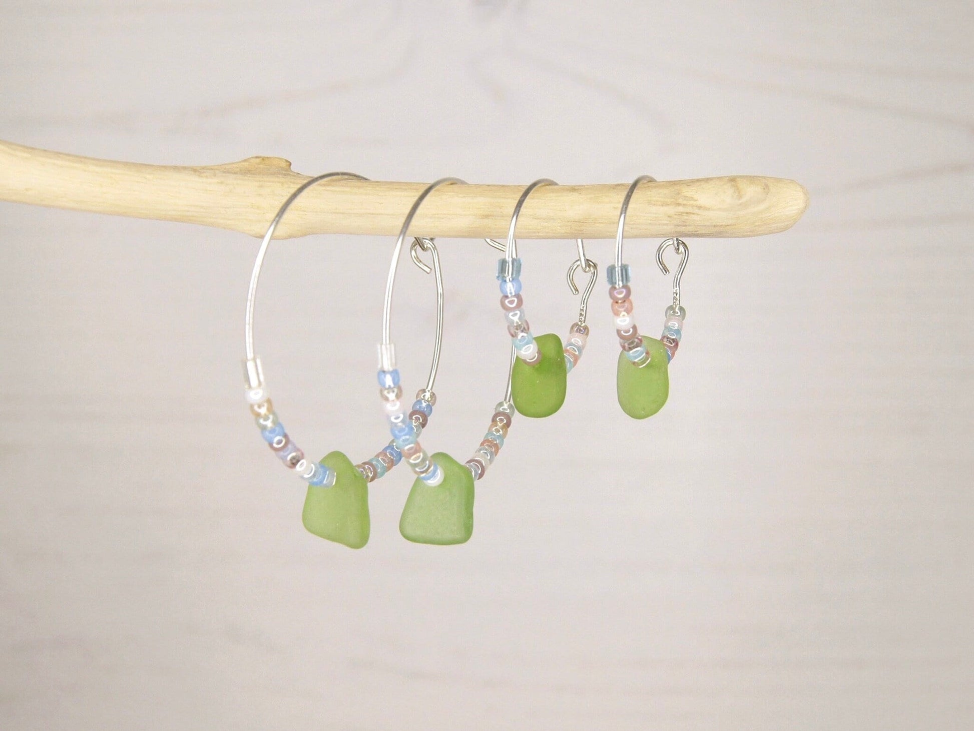 Two pairs of hoop earrings with pastel seed beads and green sea glass hanging on a driftwood against a plain background