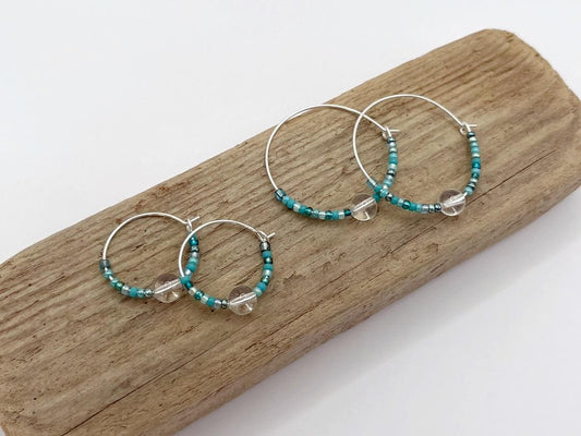 Two pairs of silver hoop earrings with turquoise seed beads and crystal laid on driftwood against a white background