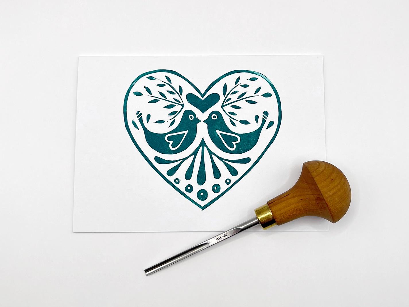 White card with an original lino printed green heart featuring two love birds on a white background. Photo includes lino cutting tool.
