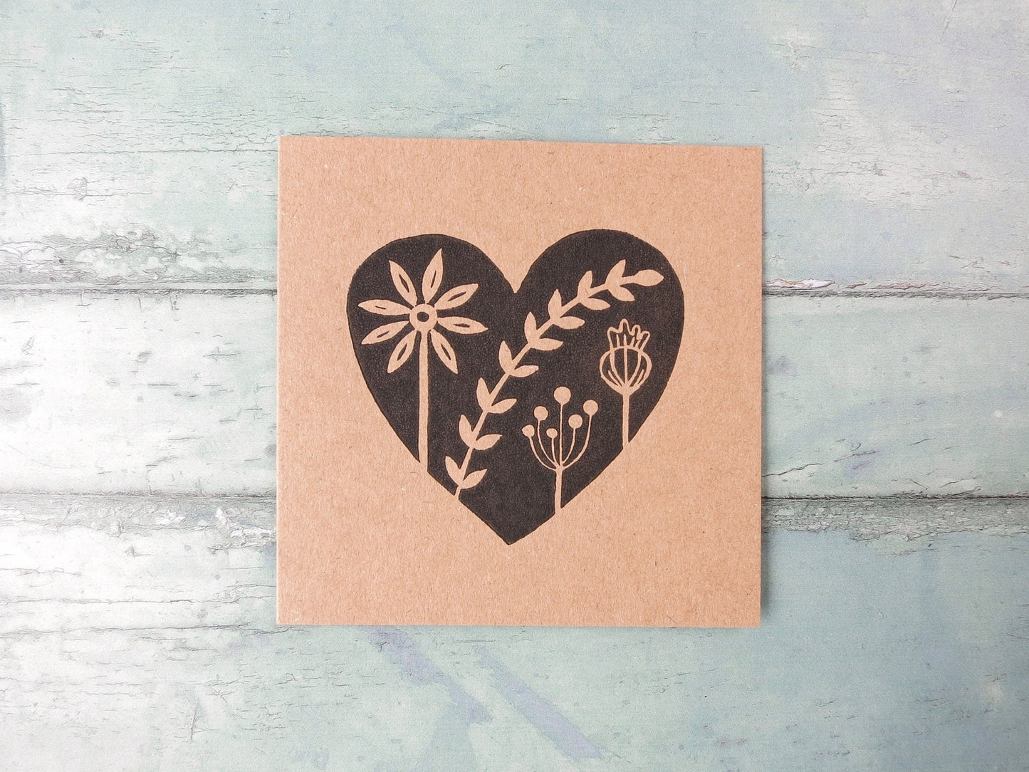 Kraft paper card with an original black lino printed heart design featuring floral elements laid on a textured surface.