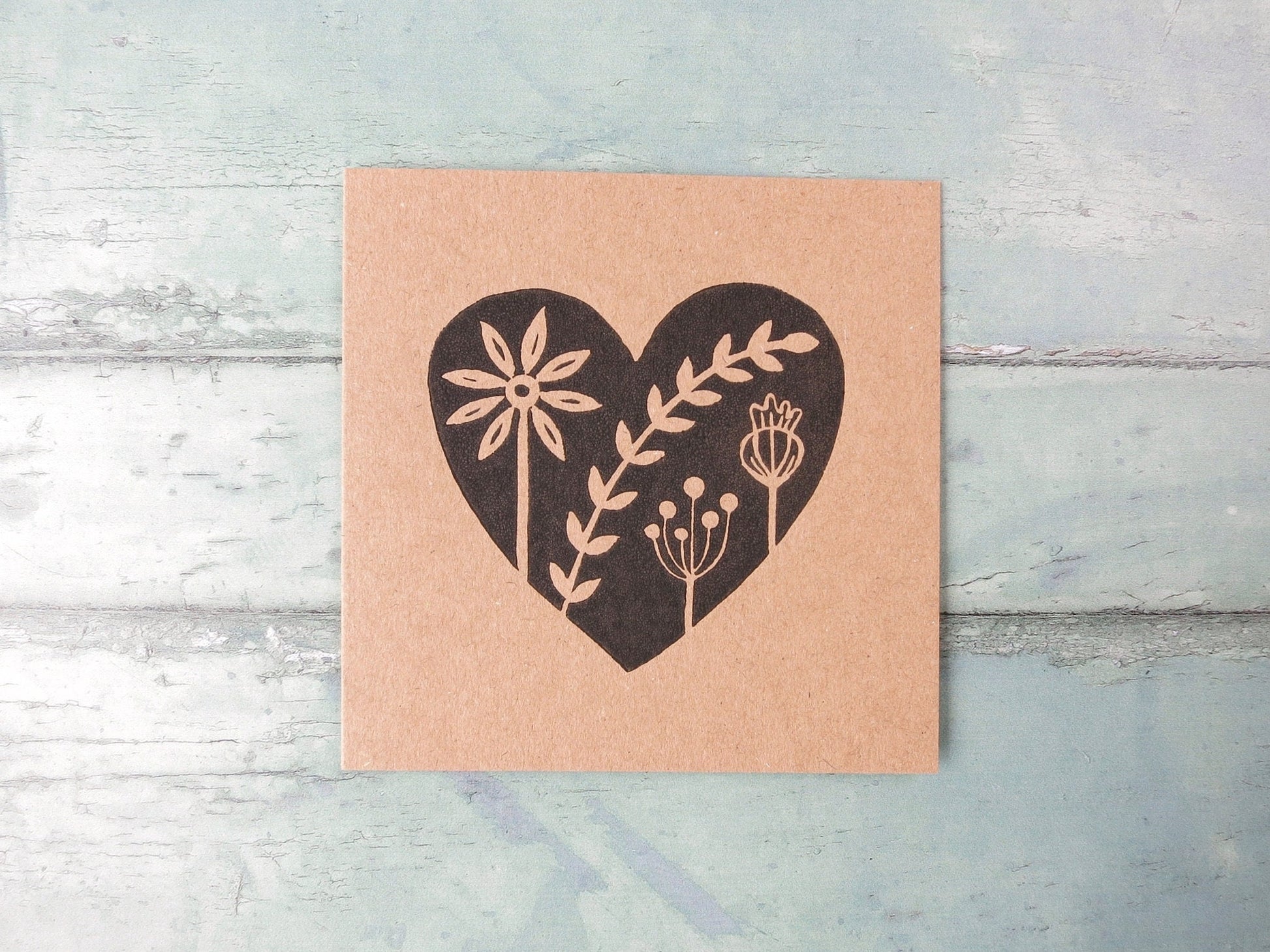 Kraft paper card with an original black lino printed heart design featuring floral elements laid on a textured surface.