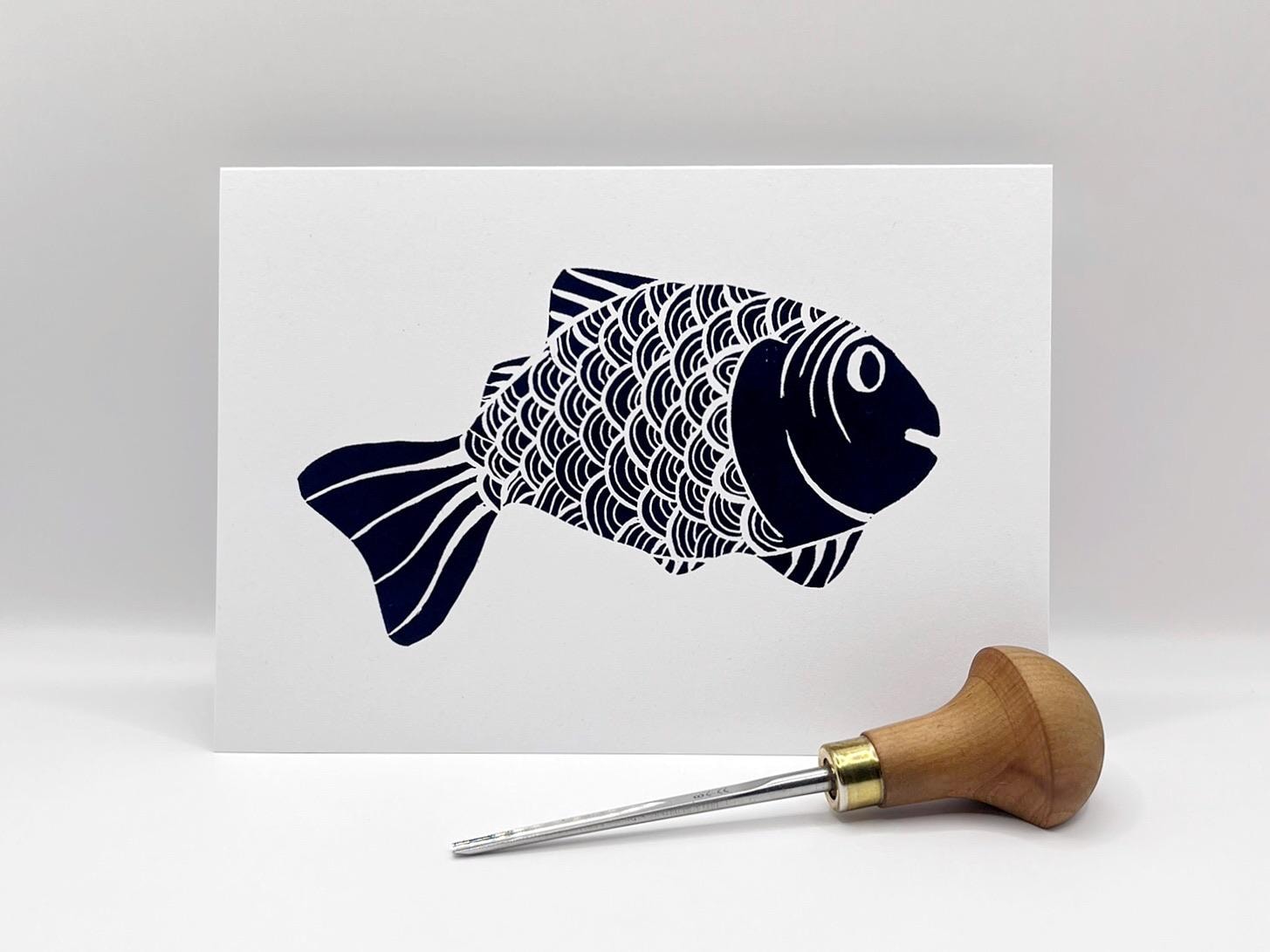 White card with an original lino printed blue fish, stood against a white background. Photo includes the lino cutting tool used.