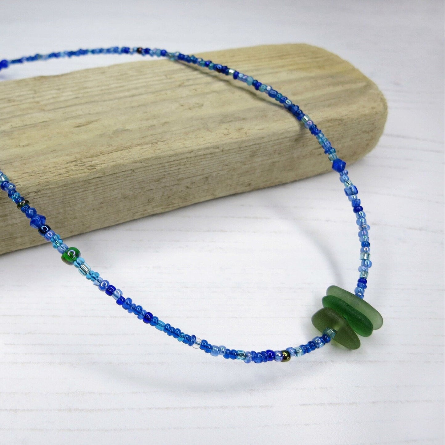 Blue seed bead necklace with green sea glass laid on driftwood against a white background