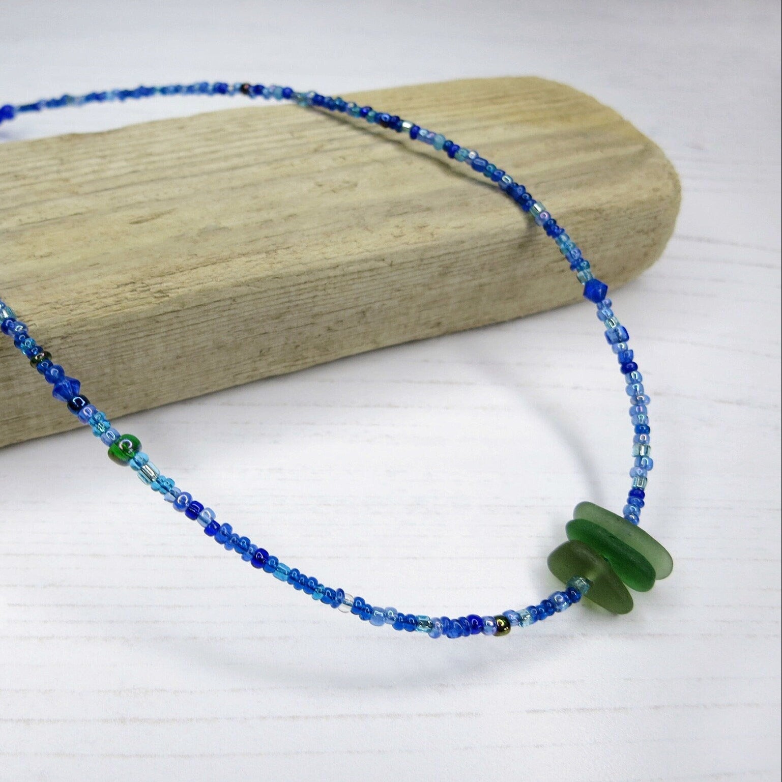 Blue seed bead necklace with green sea glass laid on driftwood against a white background