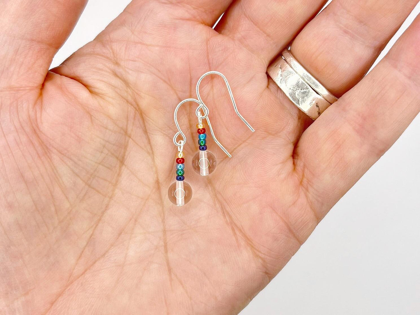 Silver earrings with clear crystal and rainbow seed beads held in a hand against a neutral background