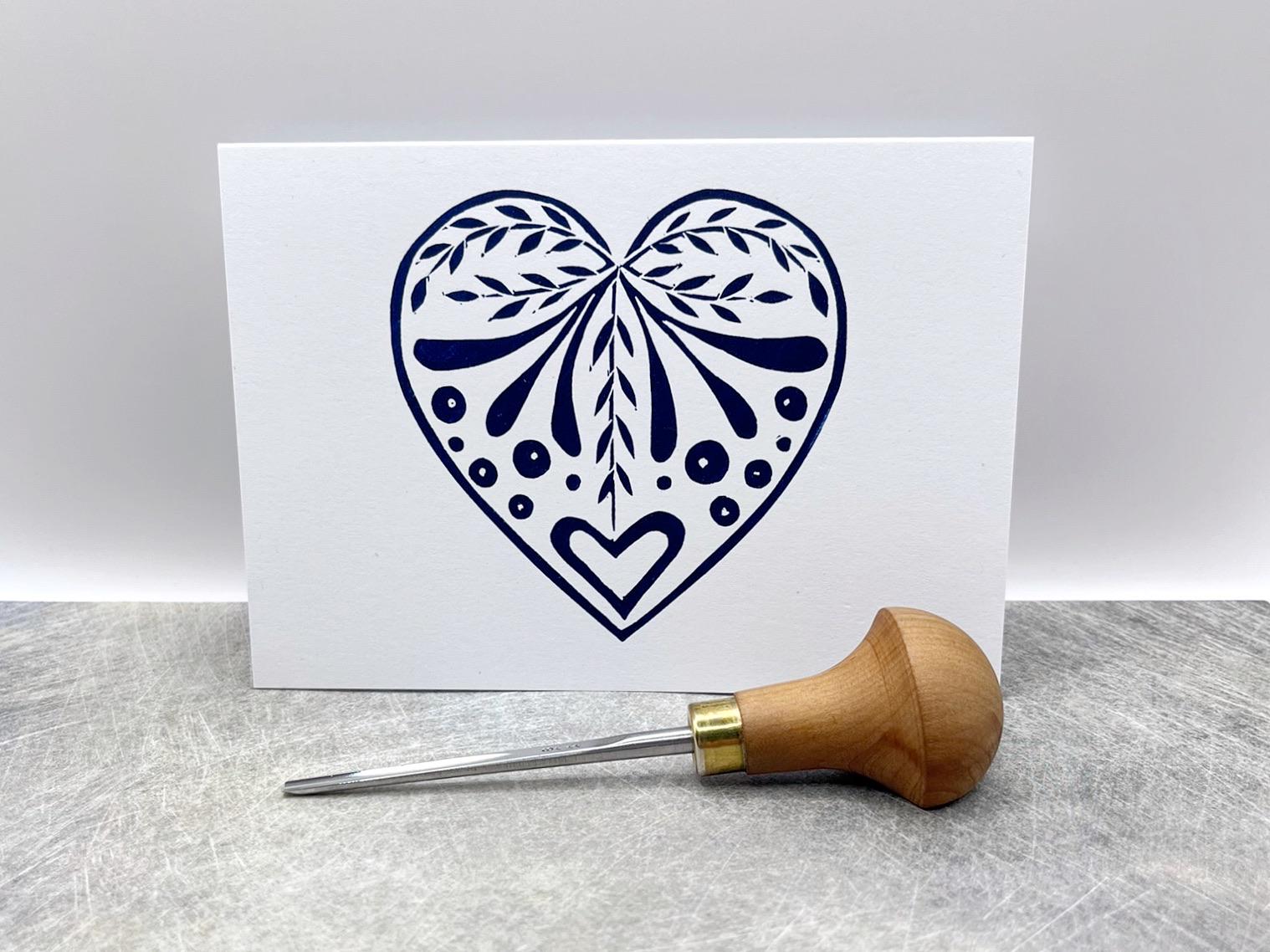 White card with an original lino printed  blue heart design stood on a textured surface. Photo includes lino cutting tool.