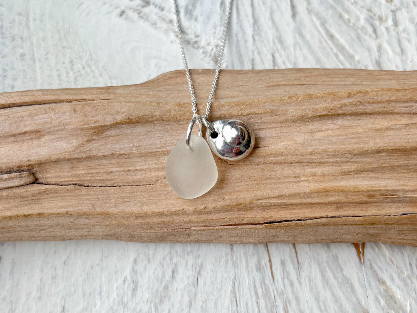 Necklace with a silver shell and white sea glass pendant laid on driftwood against a textured surface