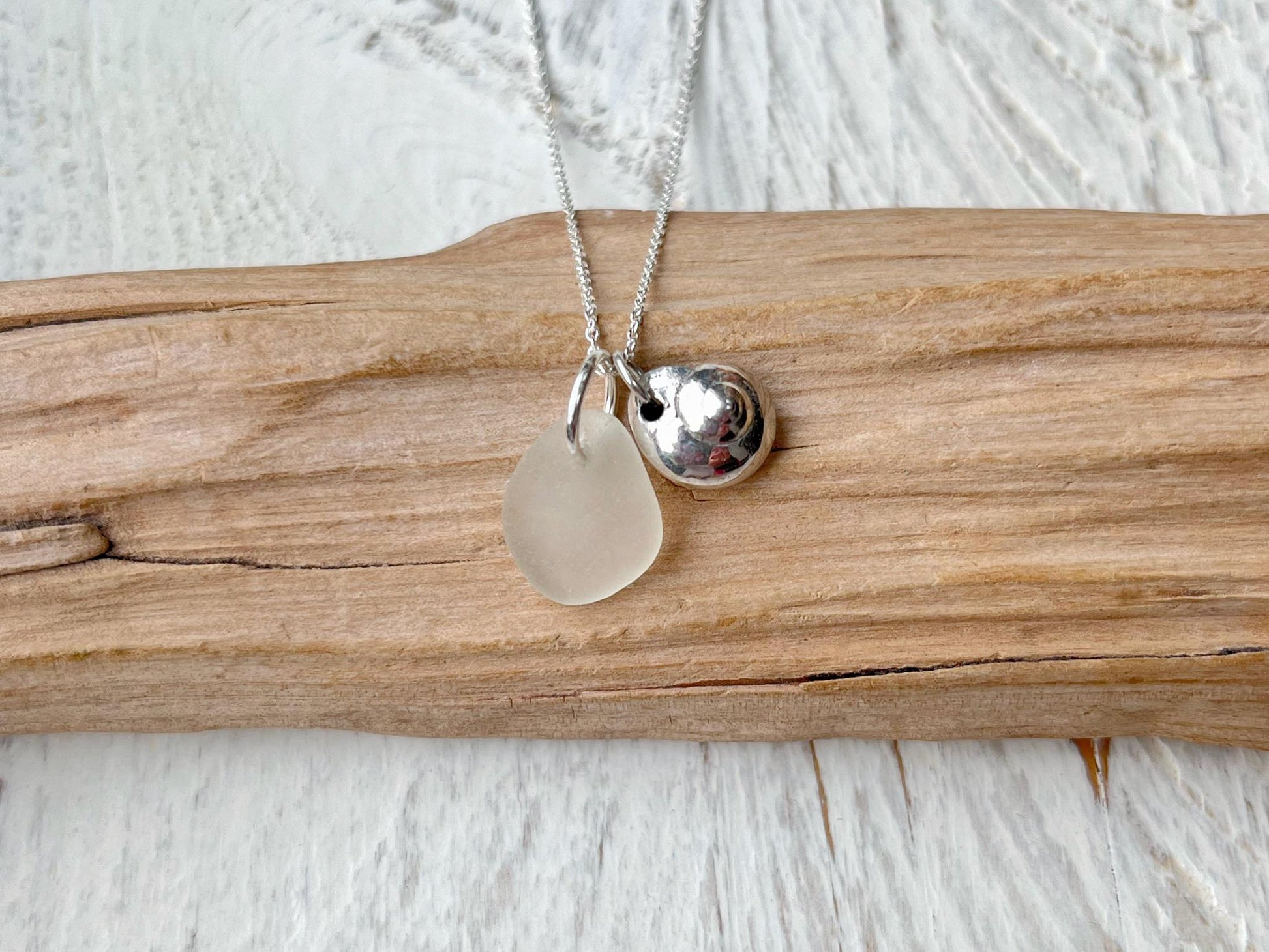 Necklace with a silver shell and white sea glass pendant laid on driftwood against a textured surface