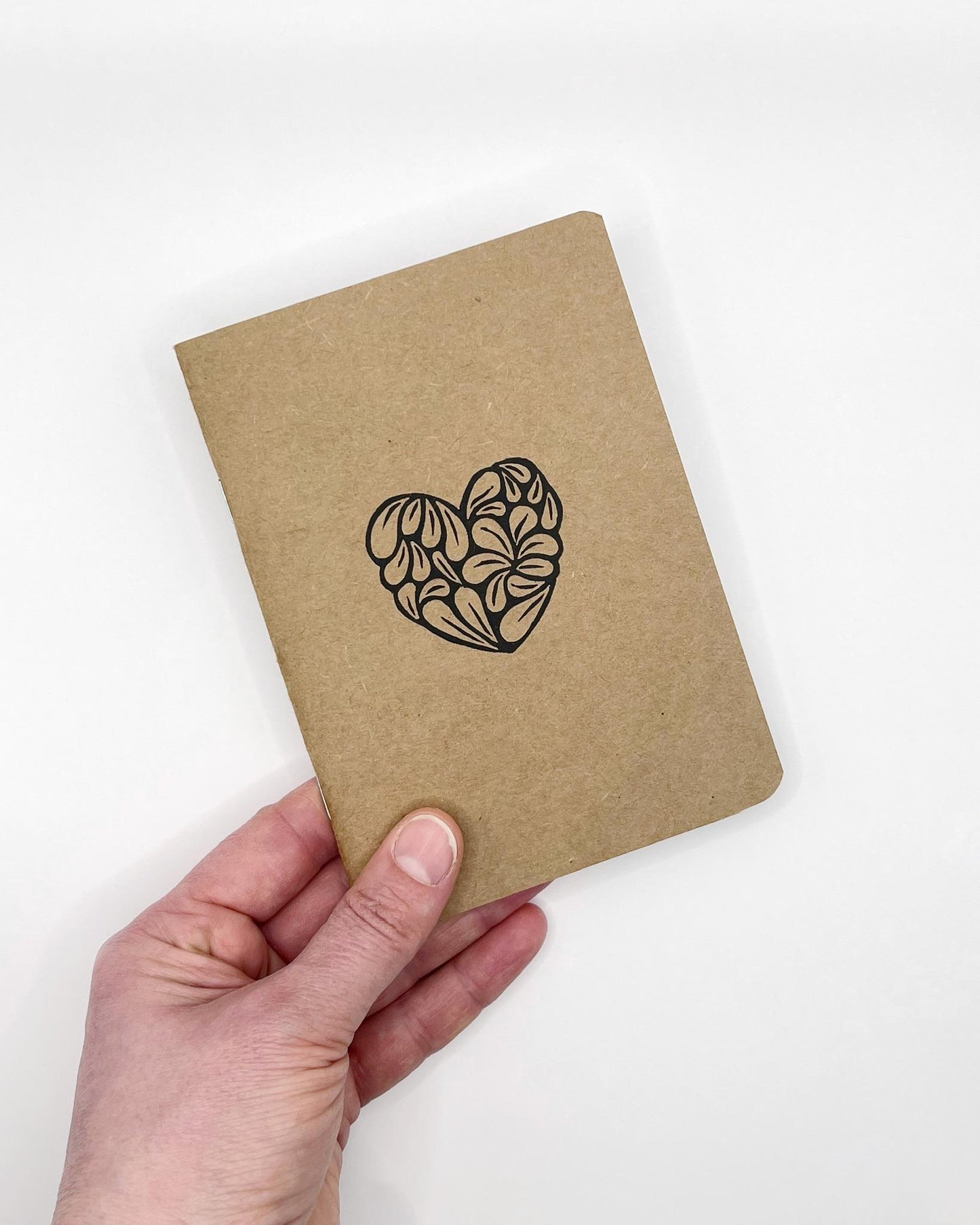 Hand holding A6 Kraft notebook with an original lino print heart. Held against a white background