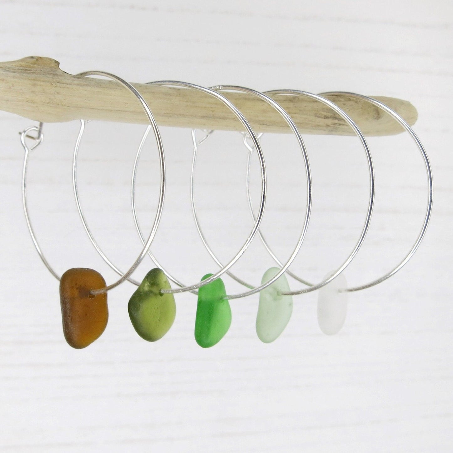 Silver hoop earrings with coloured sea glass hanging on driftwood against a white background
