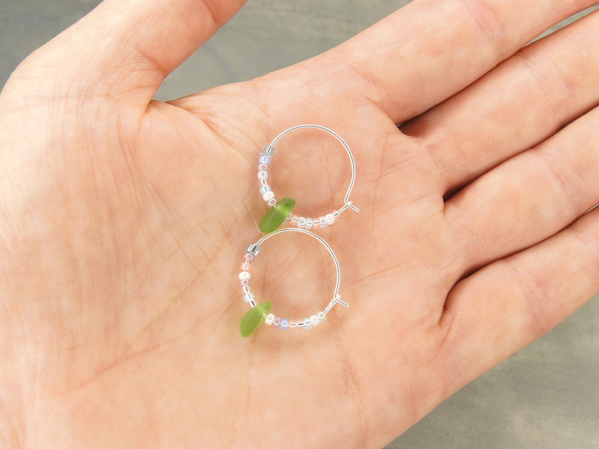 Two small beaded hoop earrings with sea glass held in a hand against a neutral background