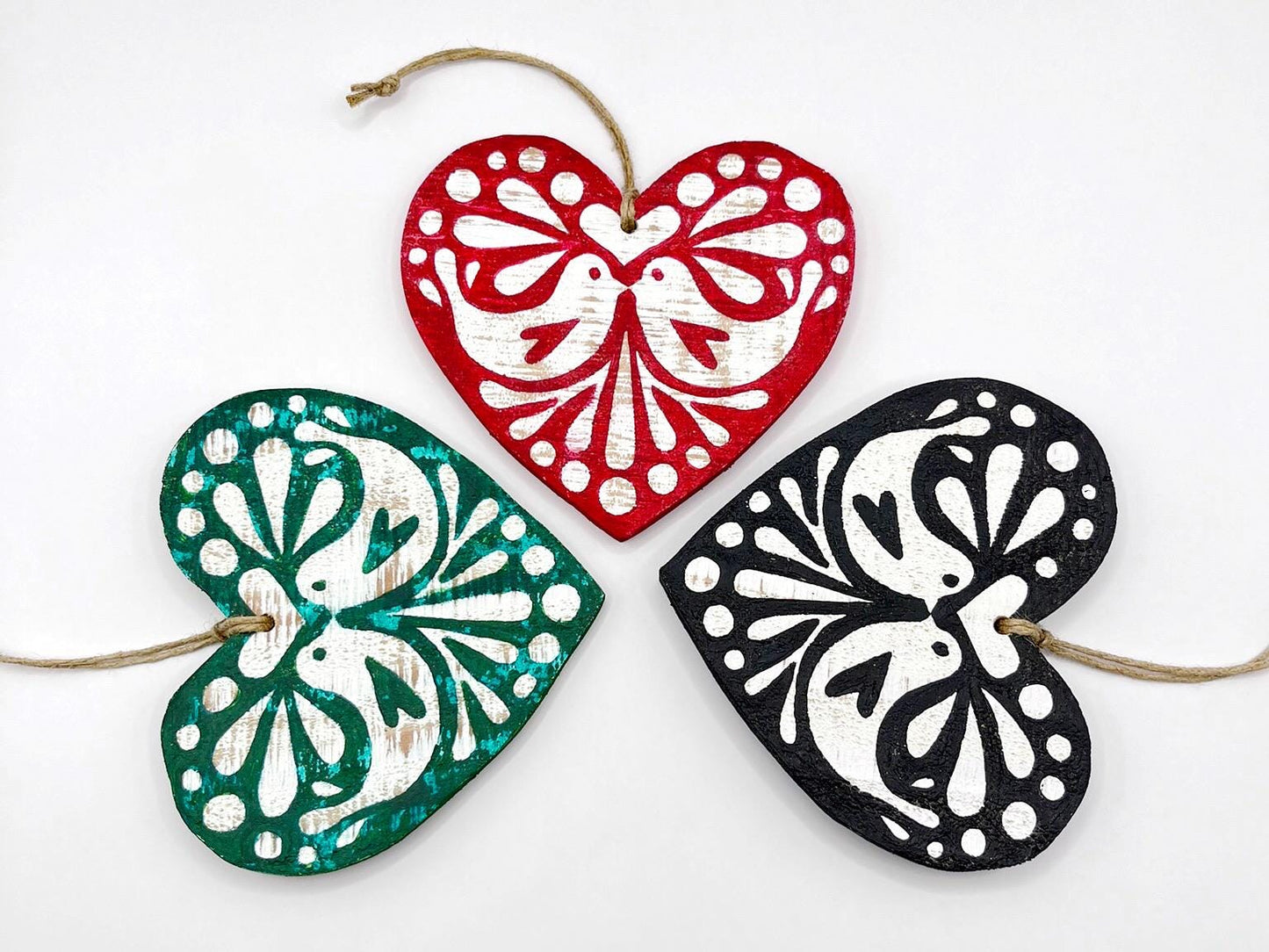 Three heart-shaped wooden hanging decorations with folk style love bird print   in red, green, and black laid on a white background