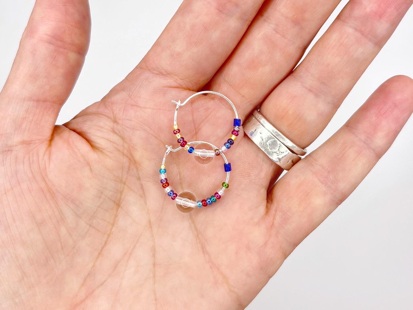 Hand holding a pair of colourful beaded hoop earrings with a plain background