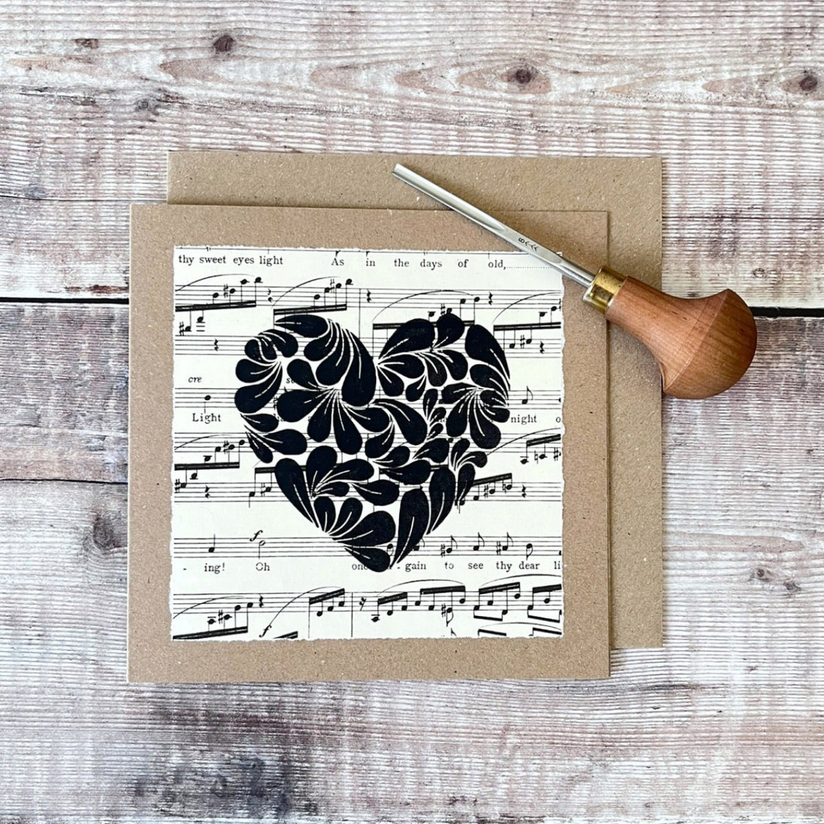 Square kraft greetings card with a lino printed heart design in black on music sheet paper mounted on the card. Laid on a matching envelope on a wooden background. Photo includes the lino cutting tool used.