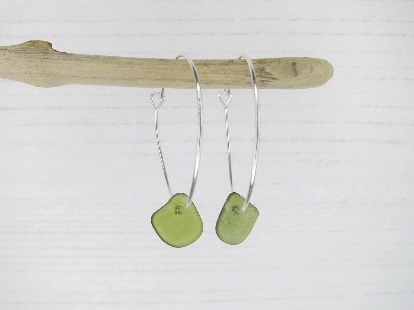 Silver hoop earrings with green sea glass hanging on driftwood against a white background
