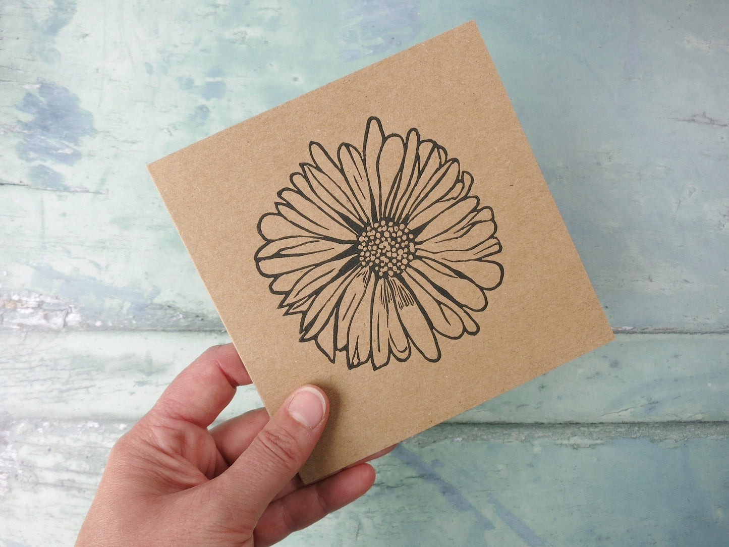 hand holding Kraft paper card with an original daisy lino print against a textured surface. 