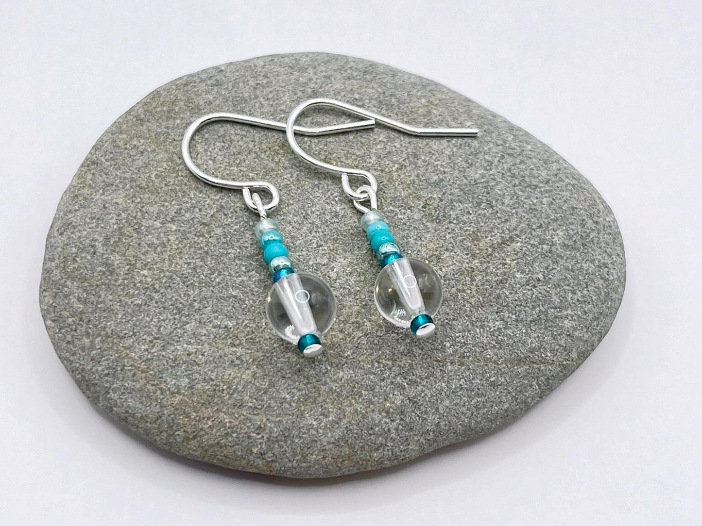 Silver earrings with clear crystal and turquoise seed beads laid on a grey pebble