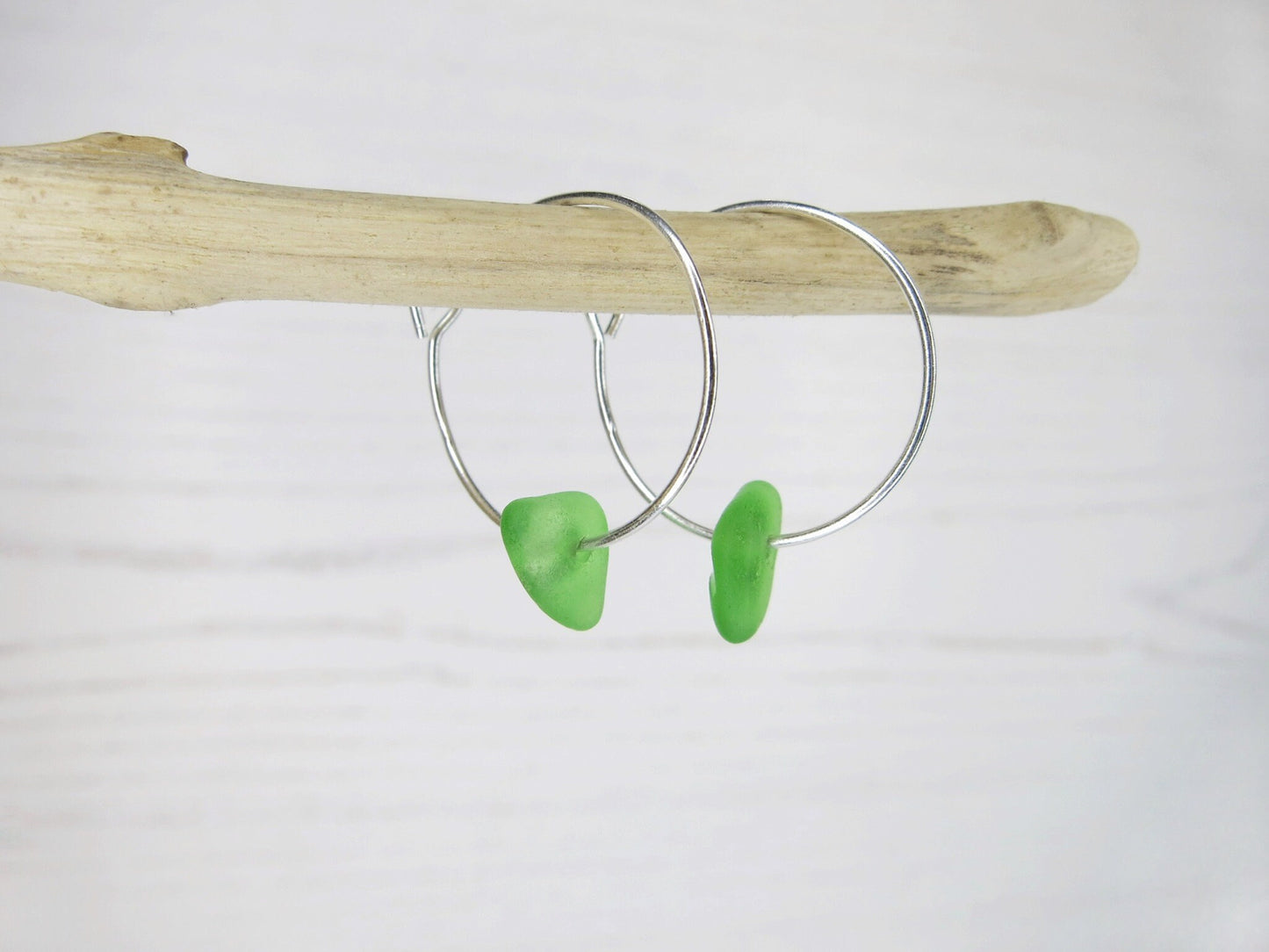 Silver hoop earrings with green sea glass hanging on driftwood against a white background