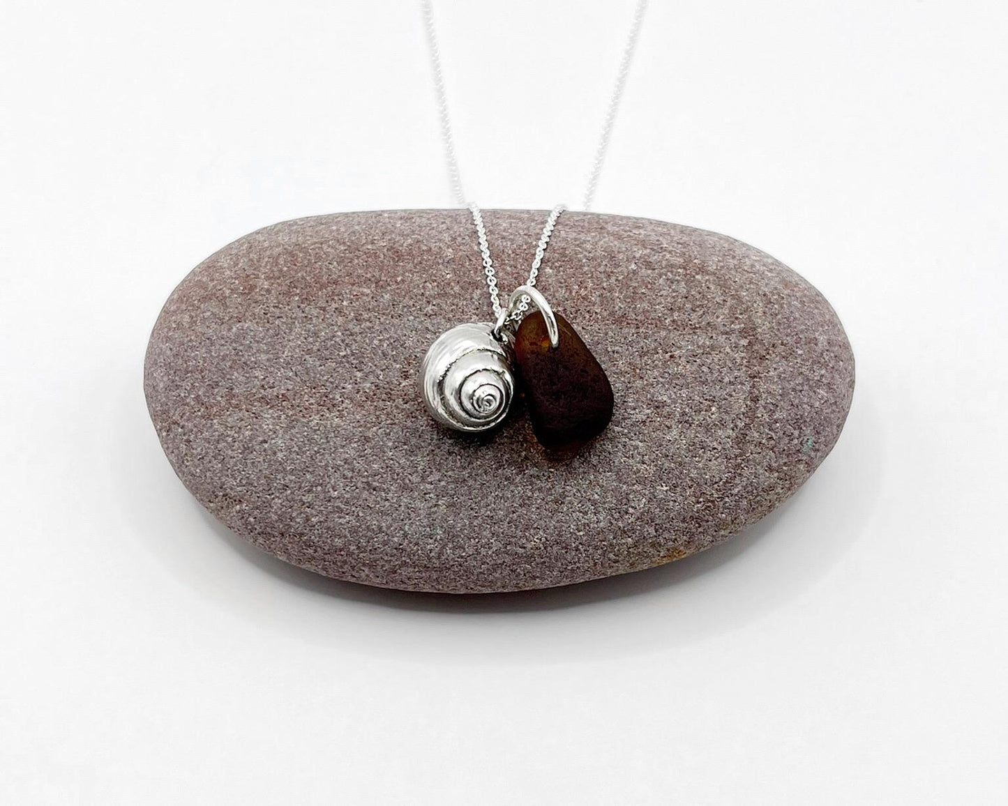 Silver necklace with burnt orange sea glass and a silver shell pendant laid on purple pebble against a white background