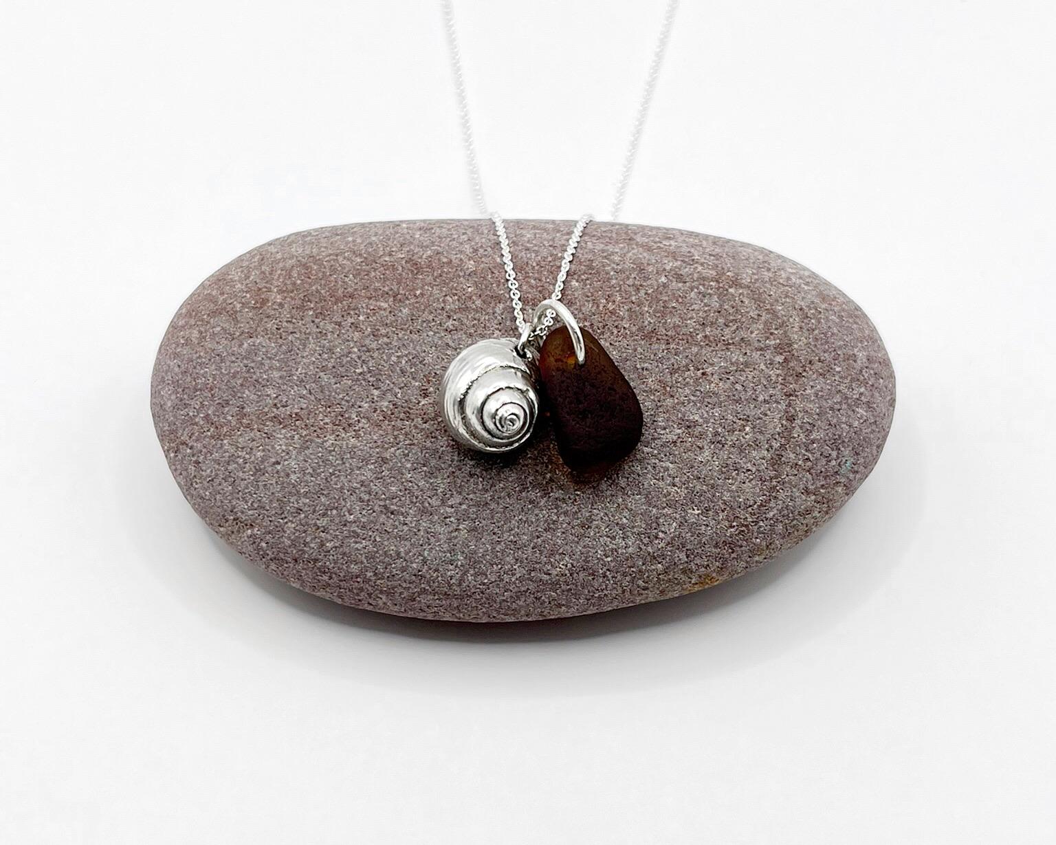 Silver necklace with burnt orange sea glass and a silver shell pendant laid on purple pebble against a white background