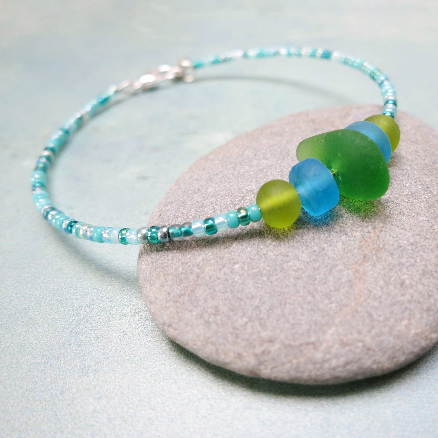Bracelet with green sea glass and turquoise and green glass beads laid on a purple pebble against a pale blue background