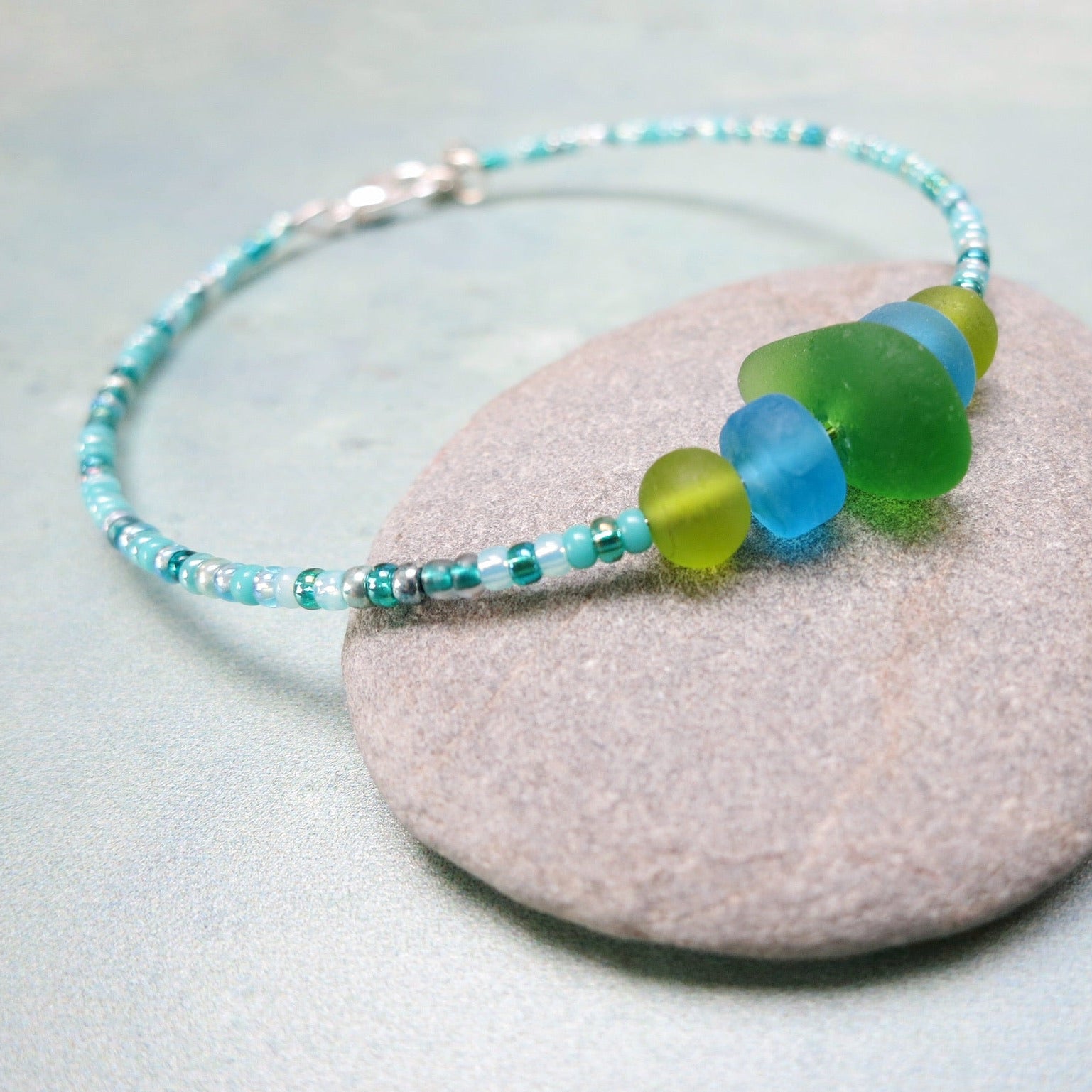Bracelet with green sea glass and turquoise and green glass beads laid on a purple pebble against a pale blue background