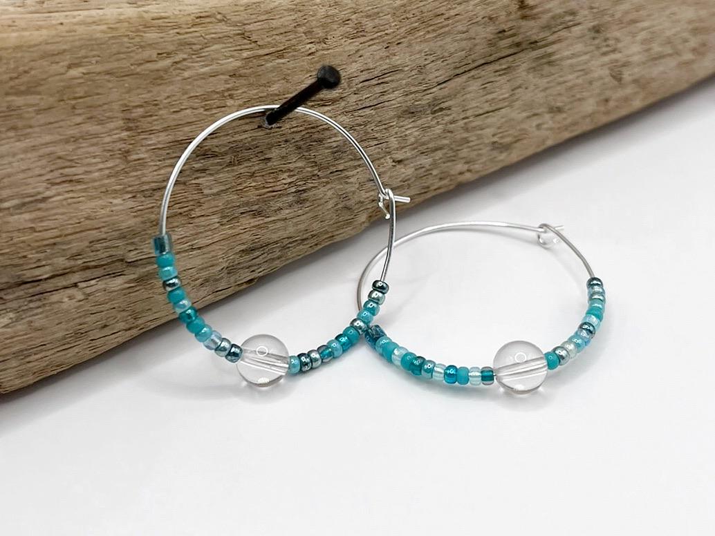 Silver hoop earrings with turquoise seed beads and crystal hanging on driftwood against a white background