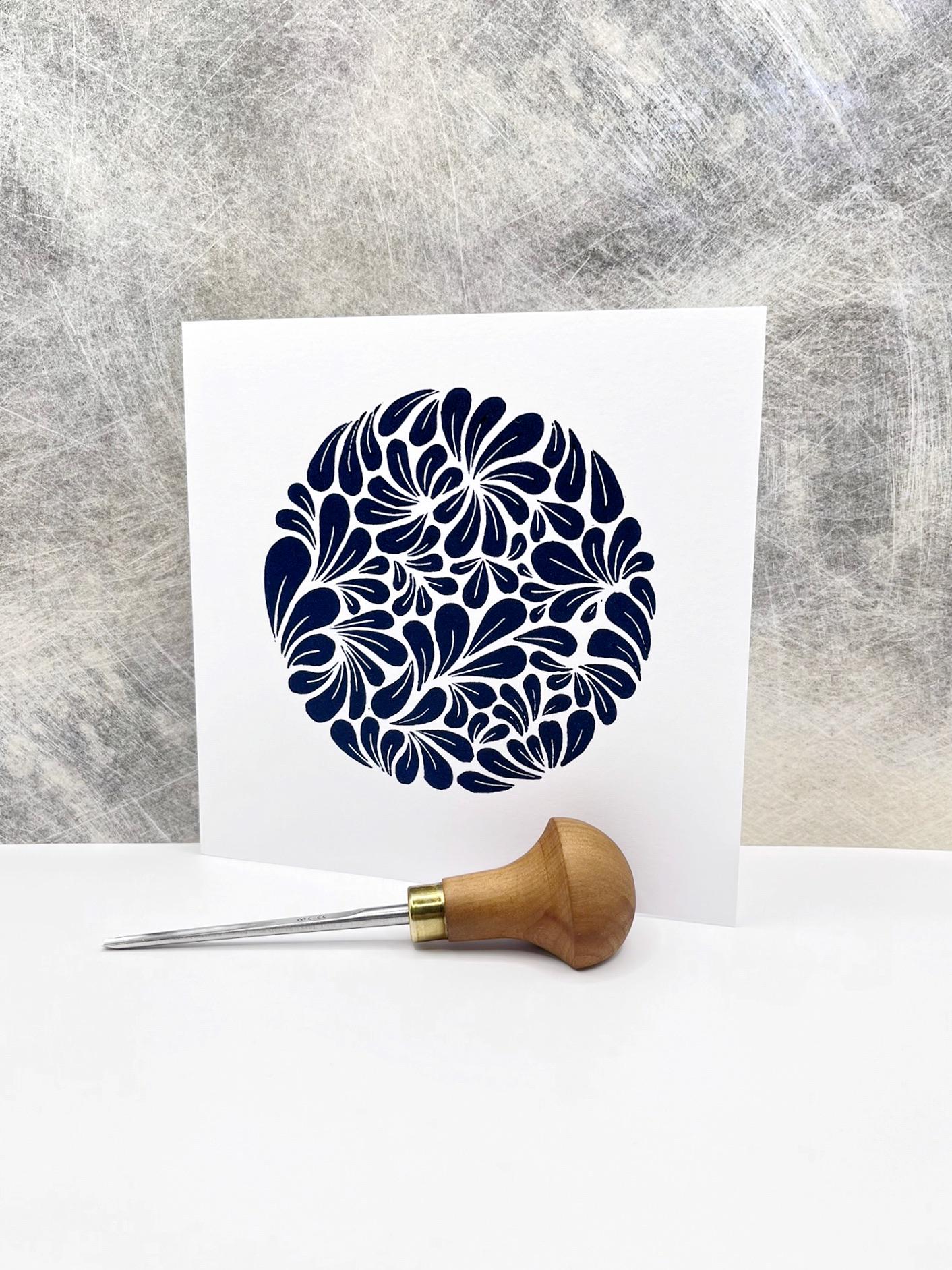 Original navy blue lino printed mussel shell pattern on a white card, stood against a textured background. Photo includes lino cutting tool