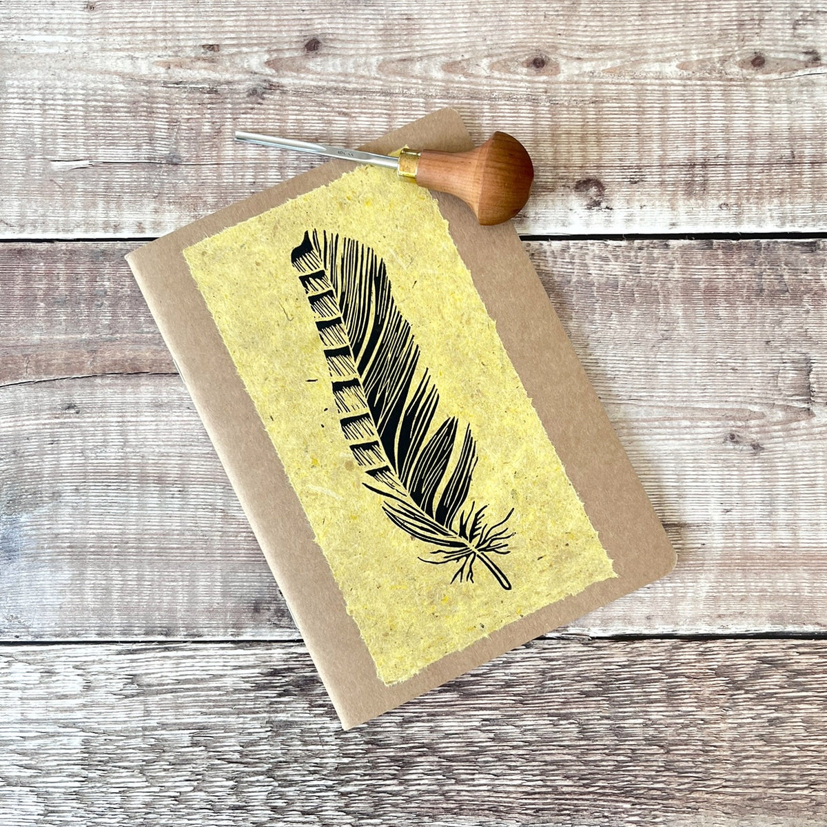 A5 kraft paper notebook or journal with a textured yellow handmade paper panel featuring a black lino ink feather illustration, photographed flat on a rustic wood plank surface. Photo includes the lino cutting tool used.
