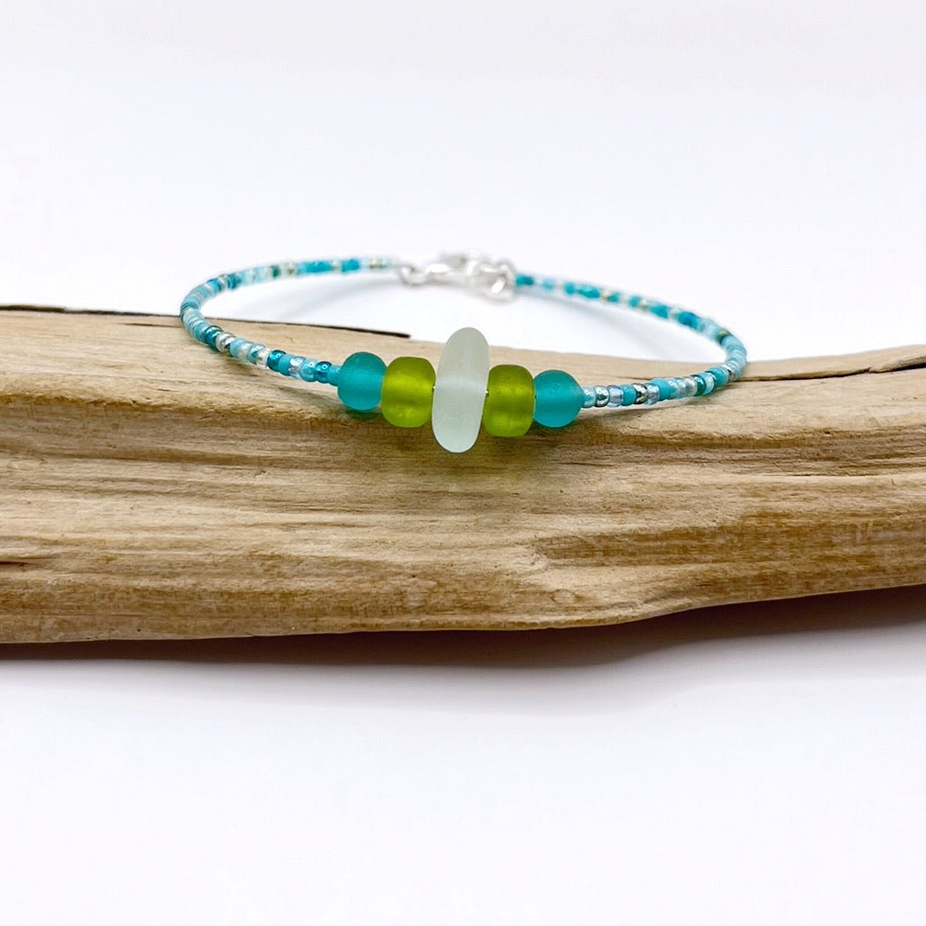 A turquoise beaded bracelet with lime and sea green frosted glass beads and a piece of sea foam coloured Cornish sea glass, finished with a silver plated lobster clasp and laid on a piece of driftwood.