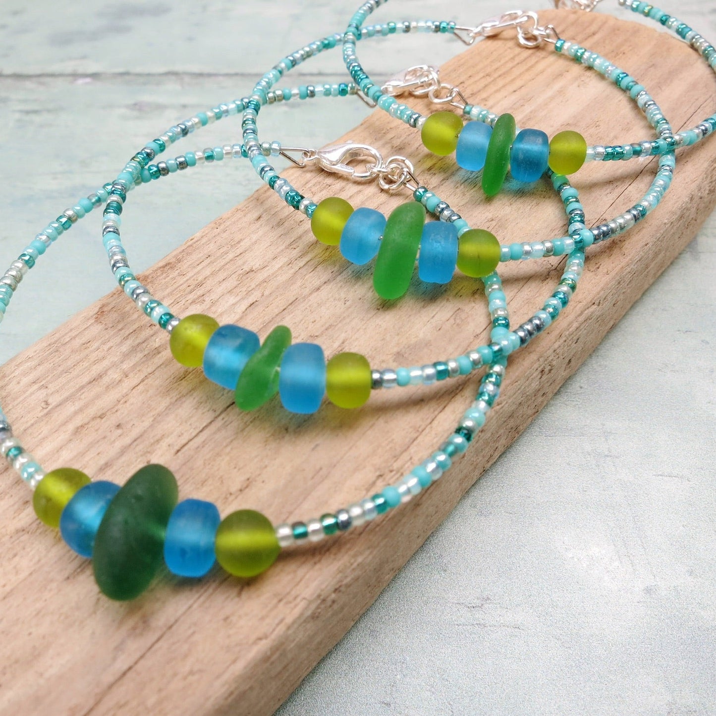 Four bracelets with green sea glass and turquoise and green glass beads laid on a driftwood against a pale blue background