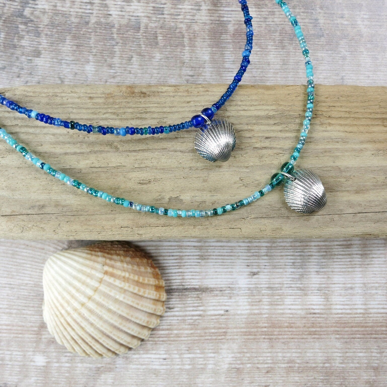 Two seed bead necklaces with silver coated shell pendants laid on driftwood against a textured surface