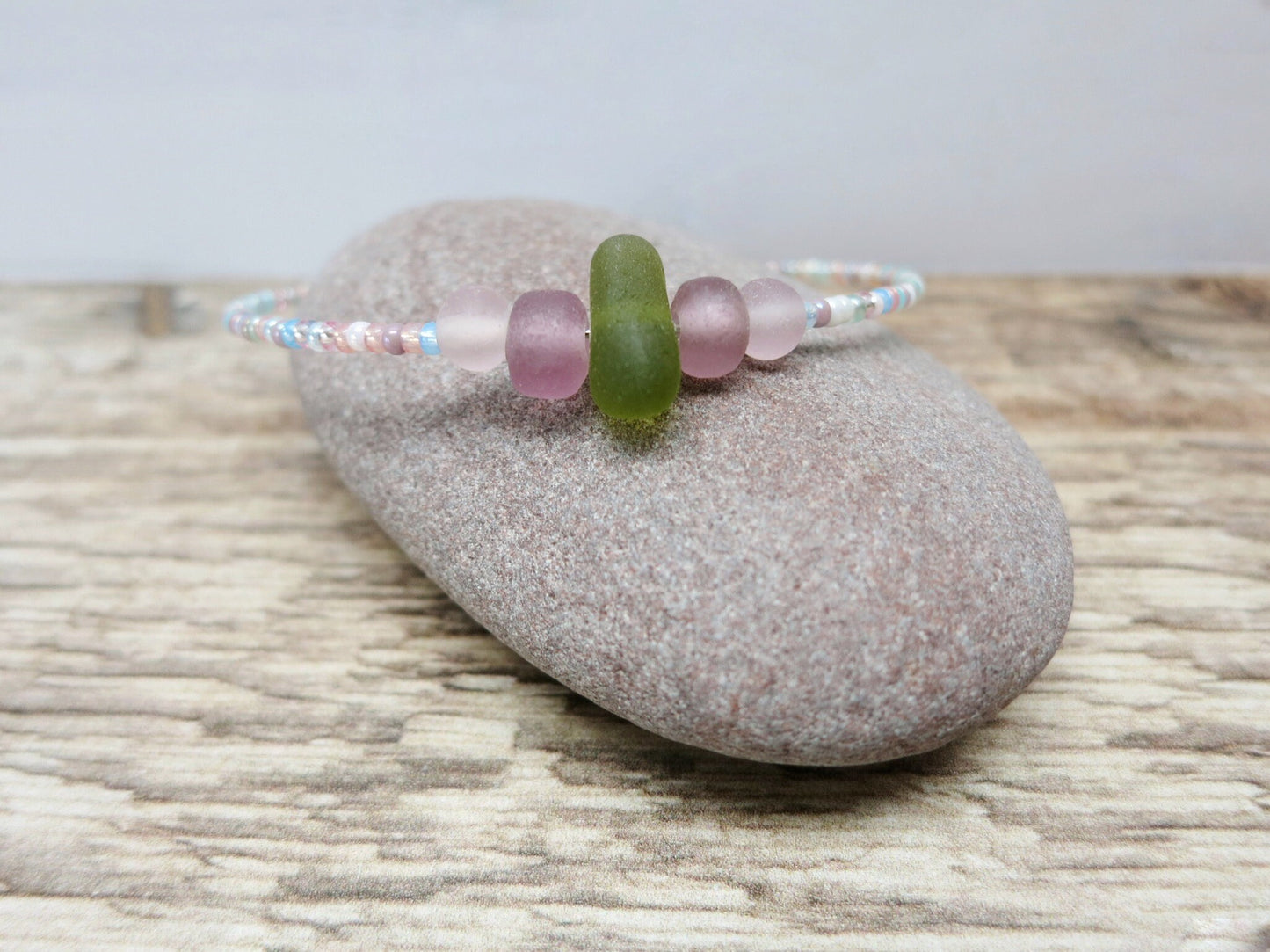 Pastel seed beaded bracelet with olive green sea glass laid on a pebble with a textured background