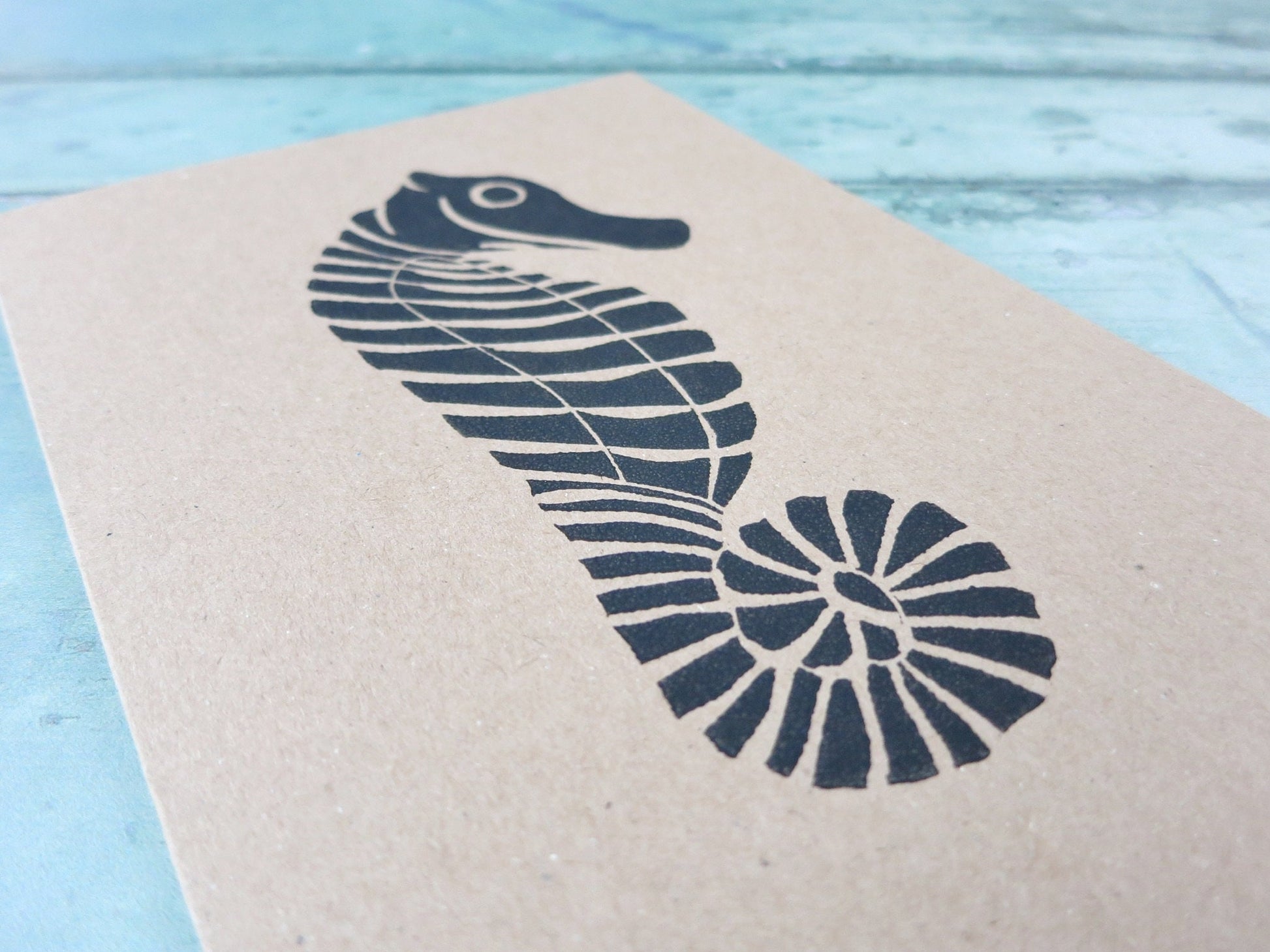 Close up of Kraft paper card with an original seahorse lino print laid on a textured blue surface. 