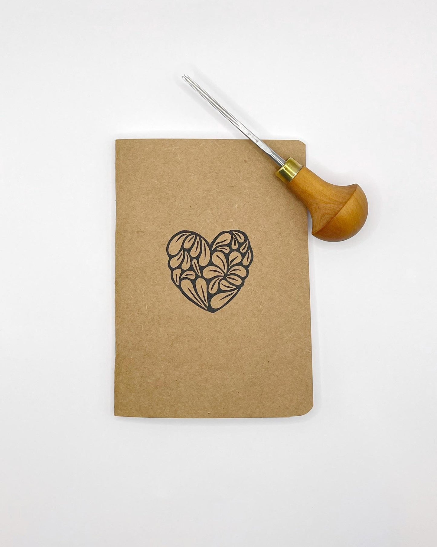 Kraft notebook with an original lino print heart design laid on a white background. Included in the photo is the lino cutting tool used.