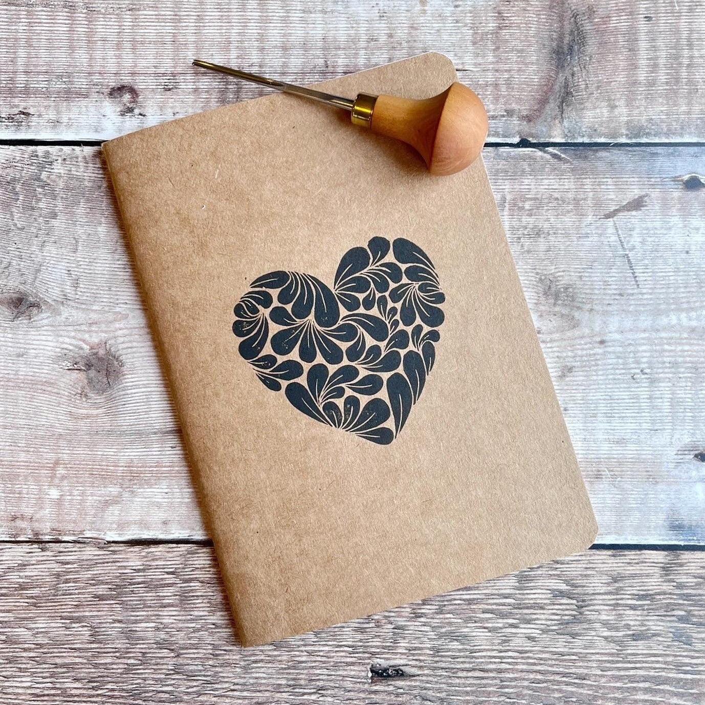 Brown notebook with original lino printed heart design on cover. Photo includes lino cutting tool used to make it laid on a textured background.
