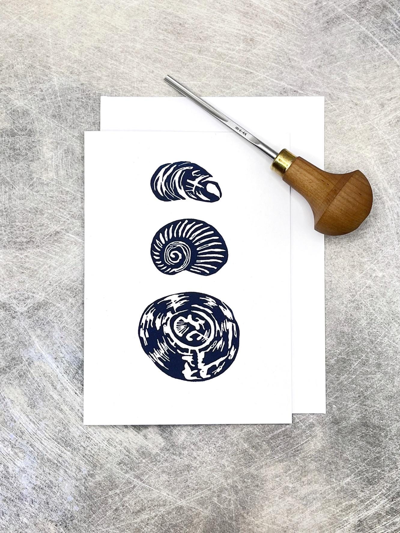 Three original lino printed shells on a white card against a grey textured background. Included in the photo is the lino cutting tool used 