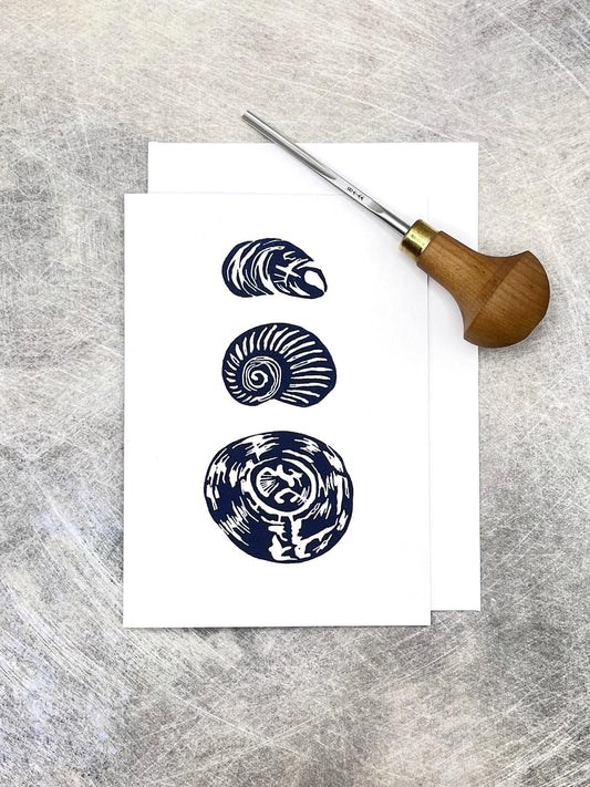 Three original lino printed shells on a white card against a grey textured background. Included in the photo is the lino cutting tool used 