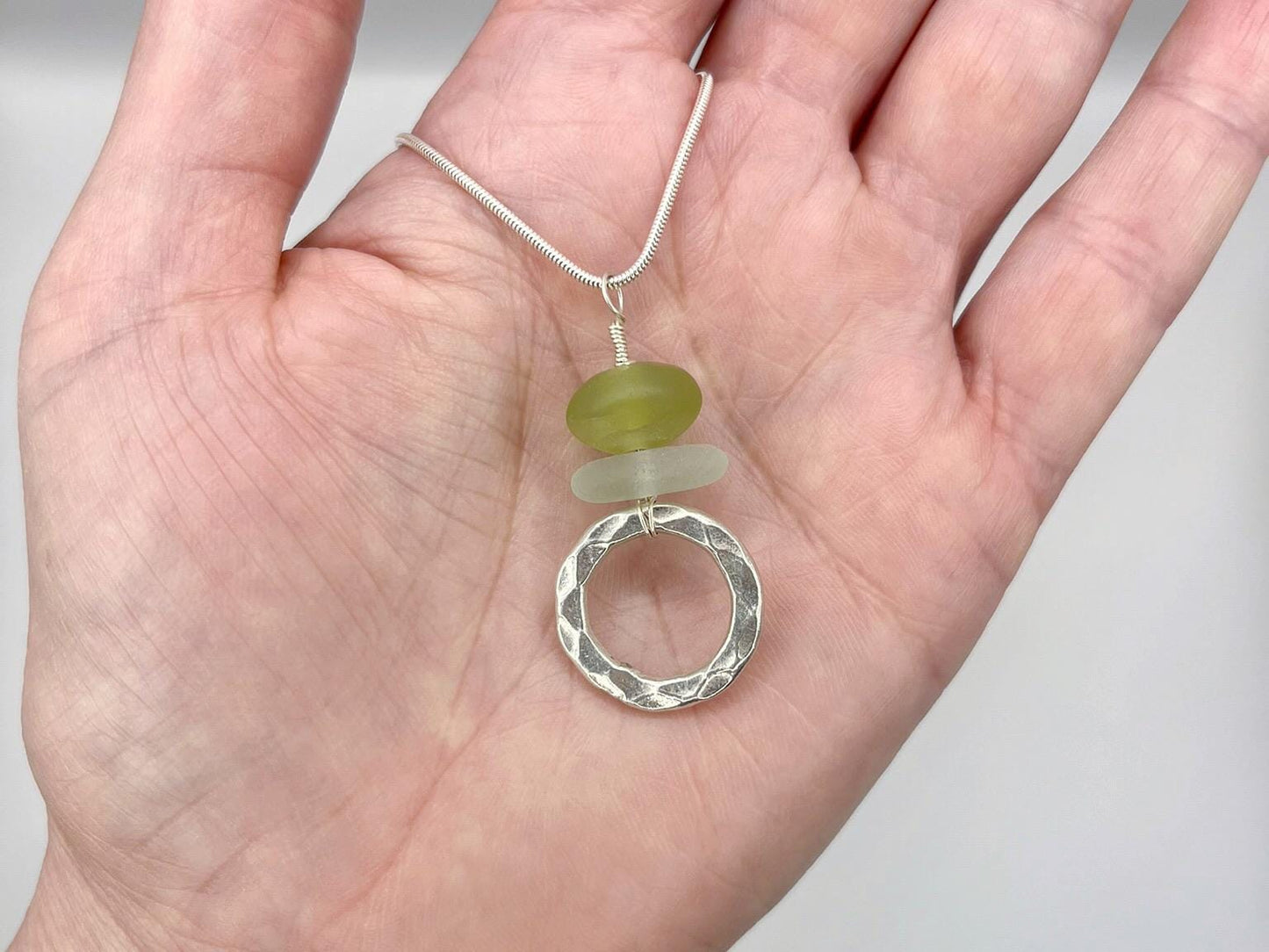Hand holding a necklace with a green jade and sea glass pendant and silver ring on a plain background
