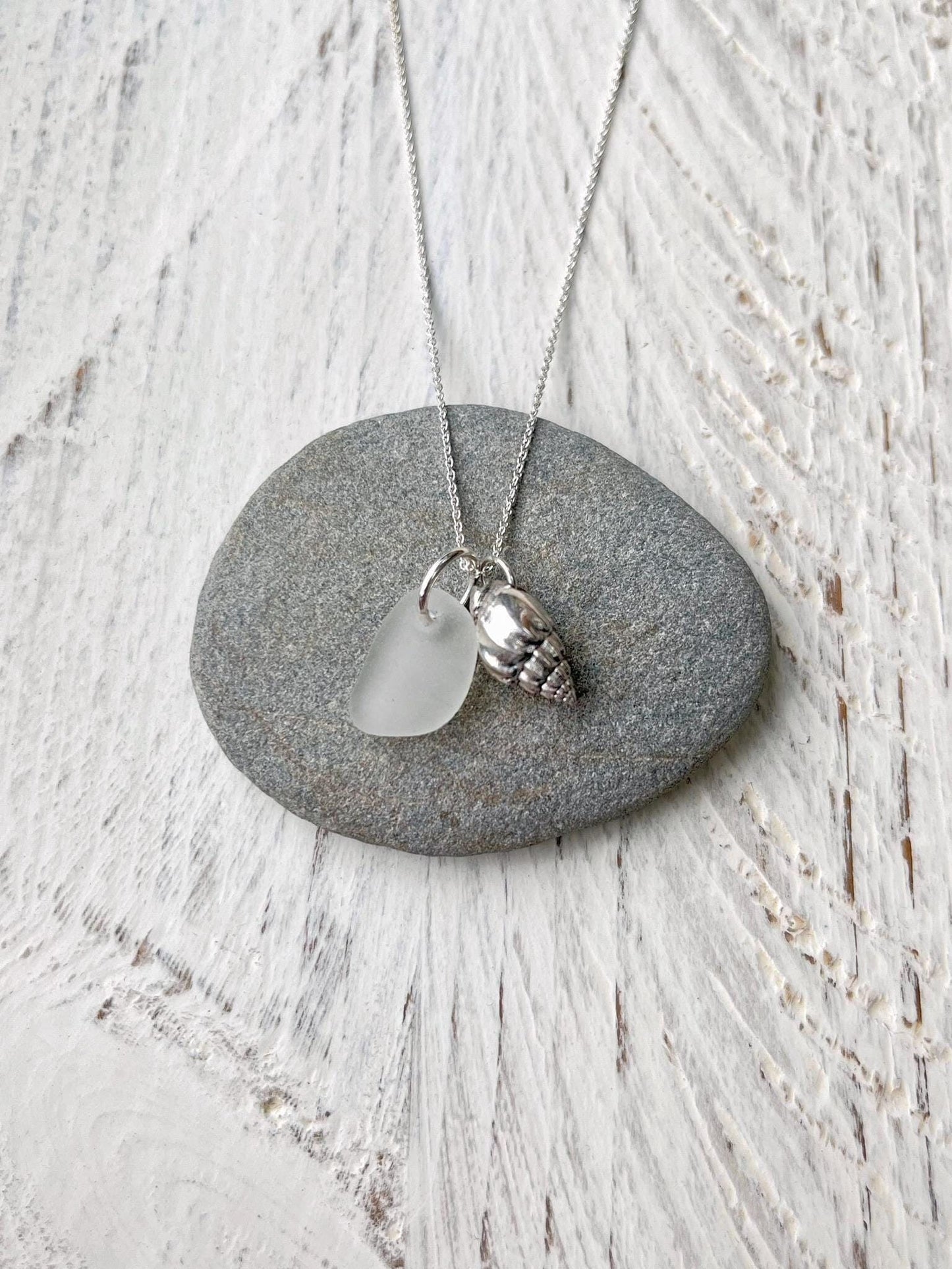 Necklace with white sea glass and silver shell pendant laid on a grey pebble against a textured surface