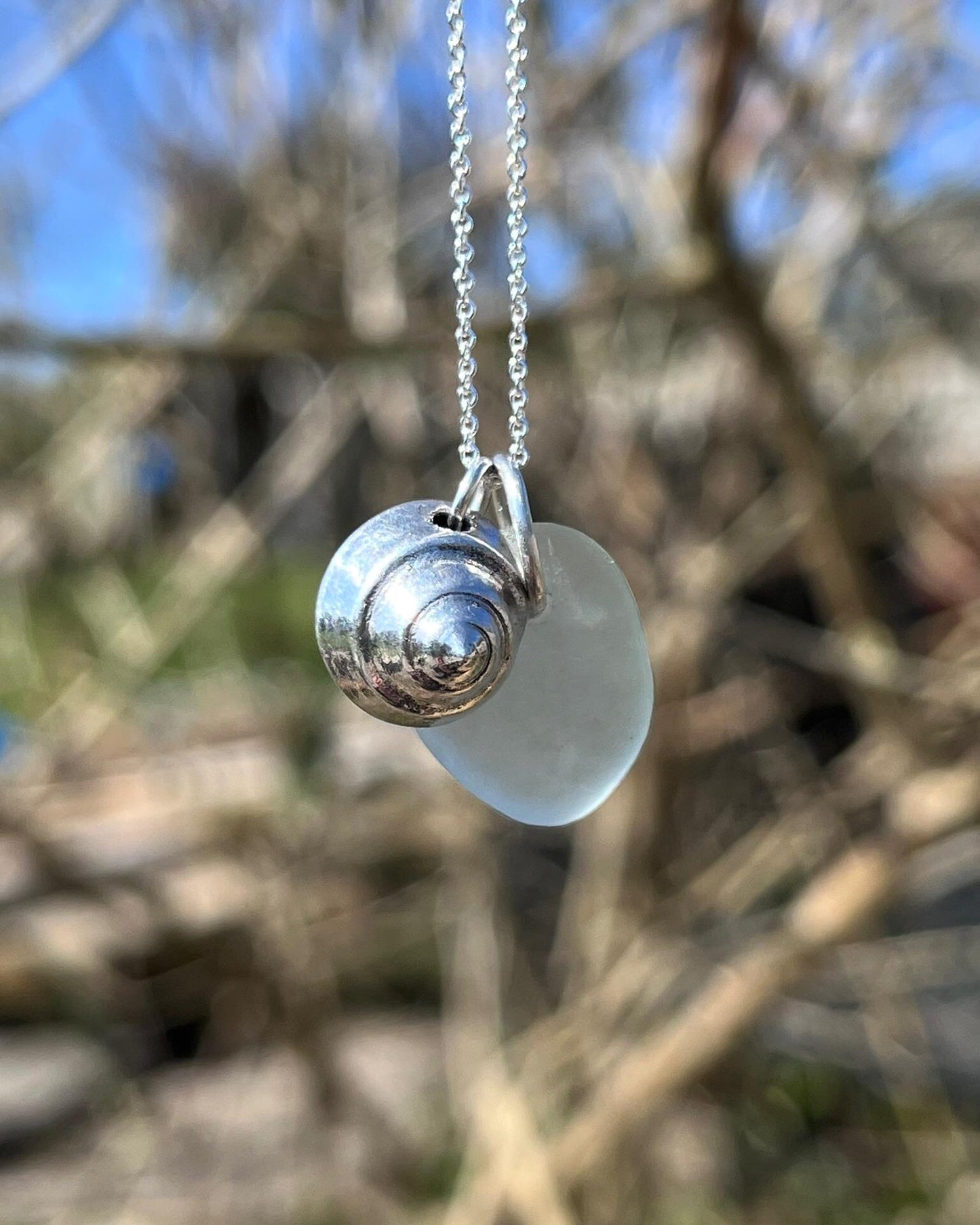 Necklace with a silver shell and seaglass pendant hanging in front of a blurred outdoor background