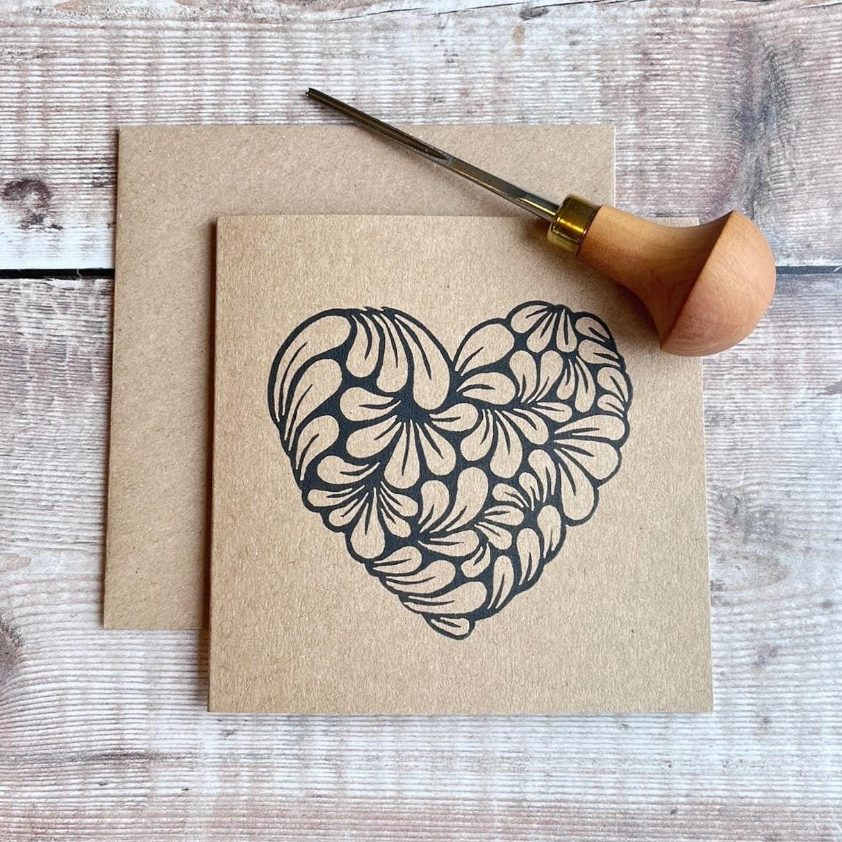 Kraft card with an original lino printed heart laid on a wooden surface. Photo includes lino cutting tool used.
