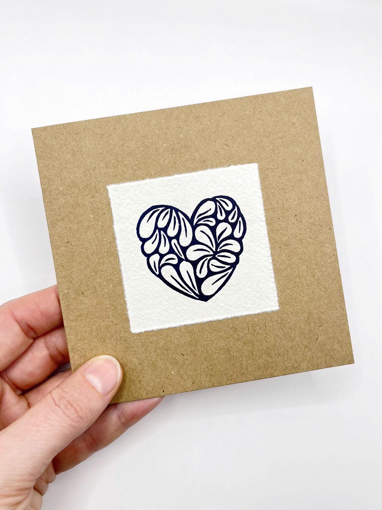 Kraft card with a white square featuring an original lino printed blue heart design held in a hand against a white background.