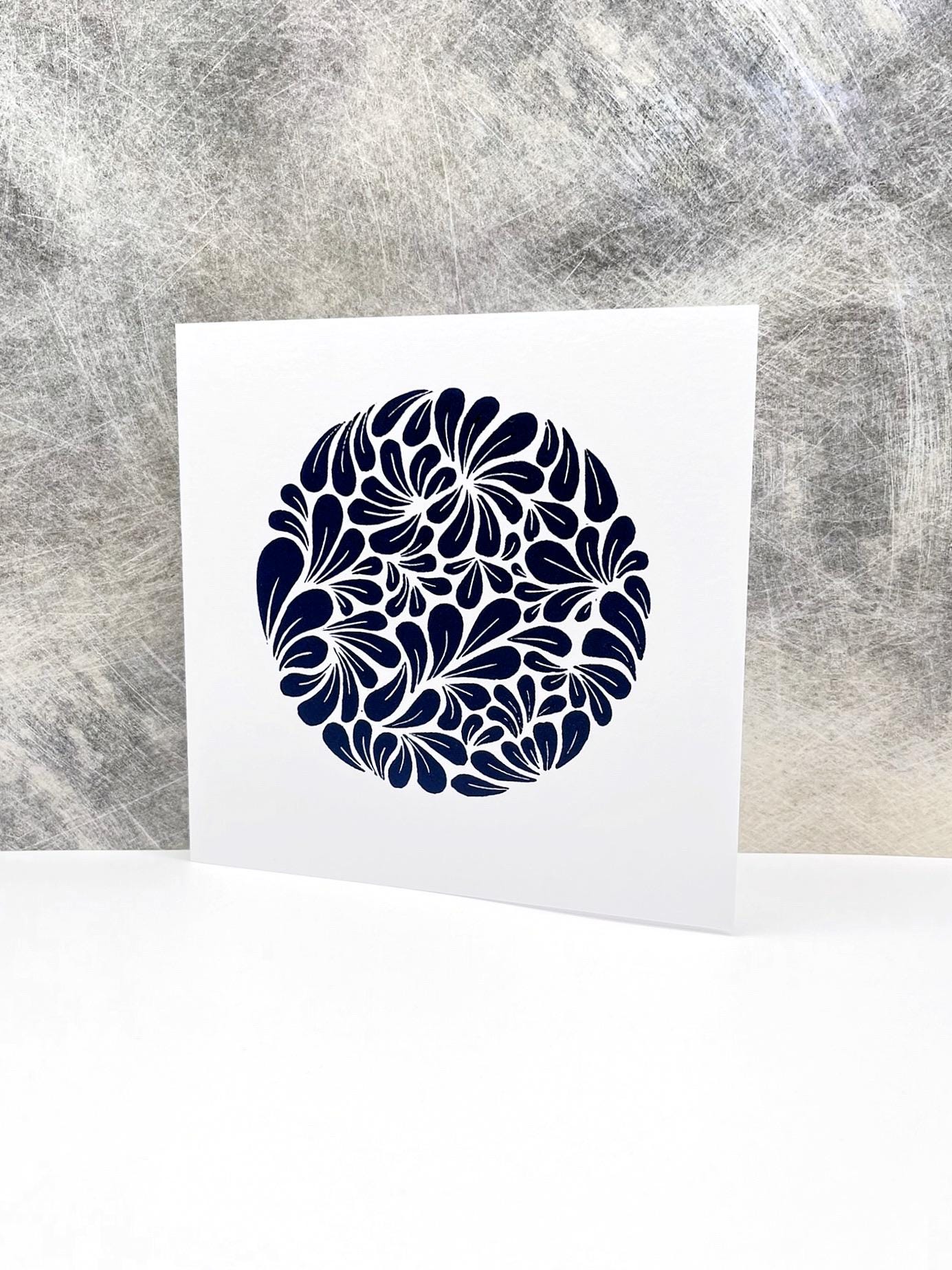 White card with a blue original lino print mussel shell pattern stood against a textured grey background