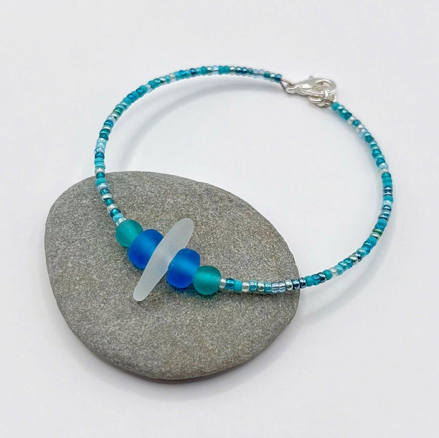 Bracelet with white sea glass and blue and turquoise beads laid on a grey pebble against a white background